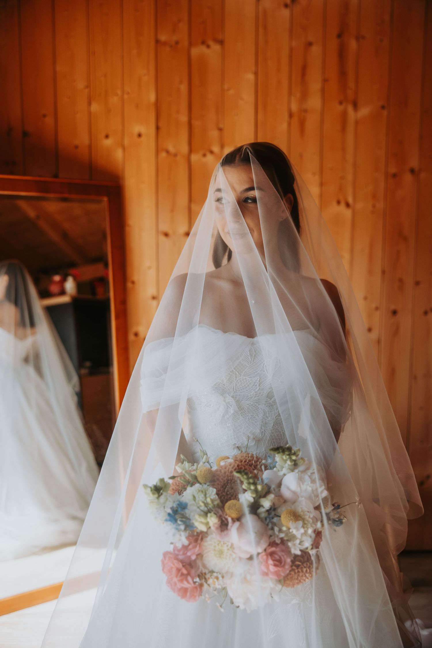 Bride in white gown holding colorful bouquet with veil draped over face in wooden room.