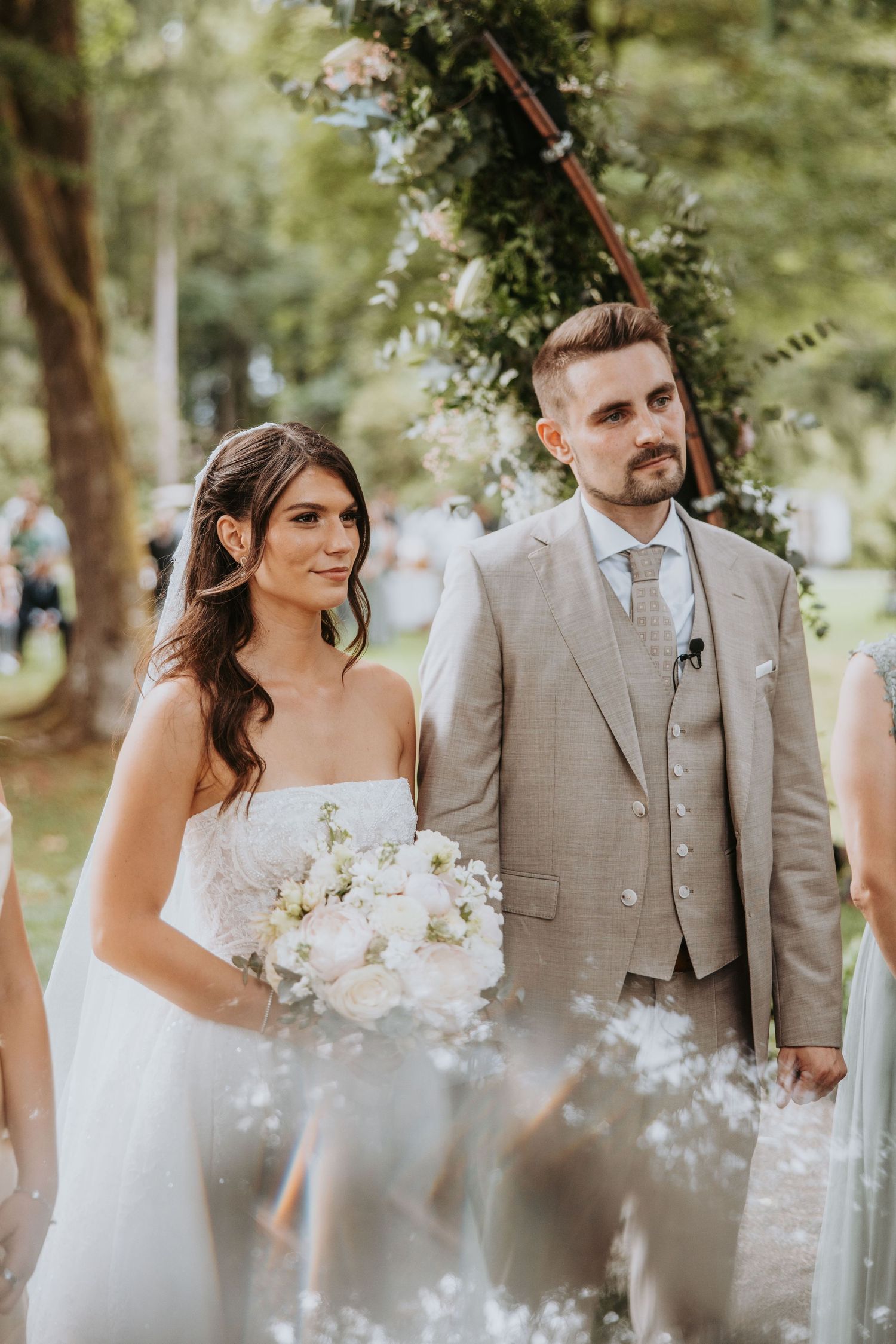 Bride in strapless white gown holding bouquet stands with groom in beige suit at outdoor wedding ceremony.