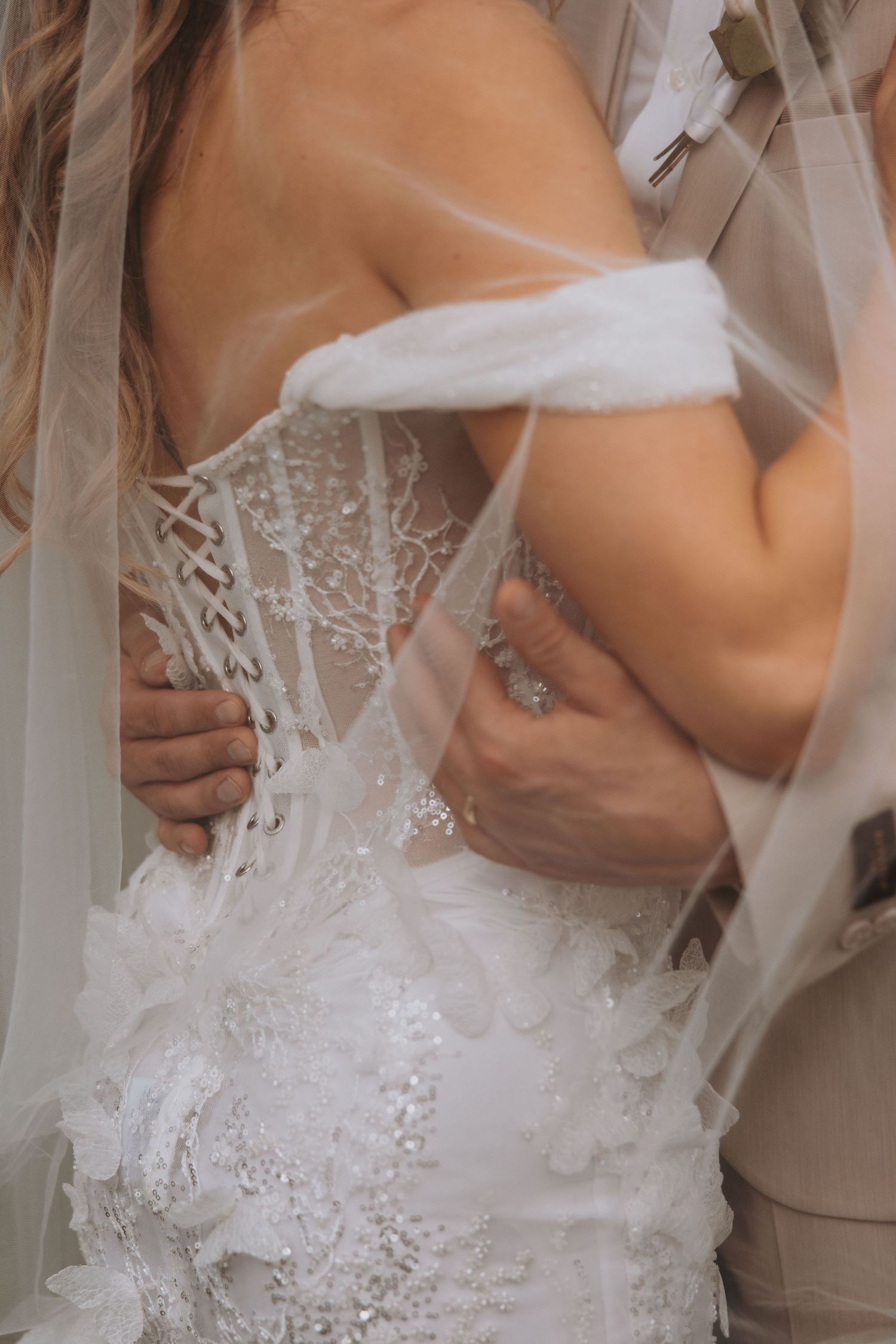 Close-up of bride in intricate lace corset gown with groom embracing her from behind under sheer veil.