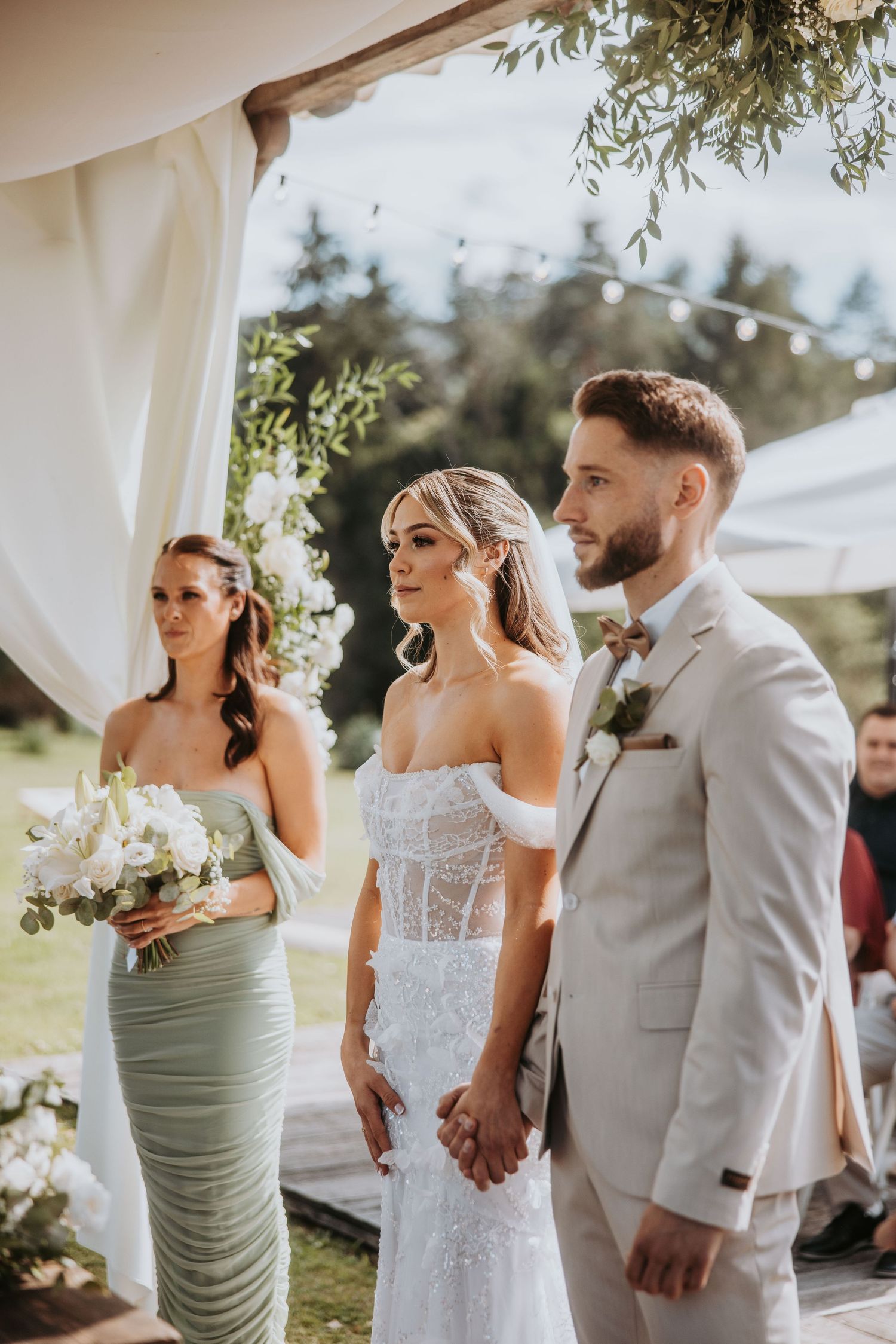 Bride and groom holding hands at outdoor wedding ceremony with bridesmaid in sage green dress.