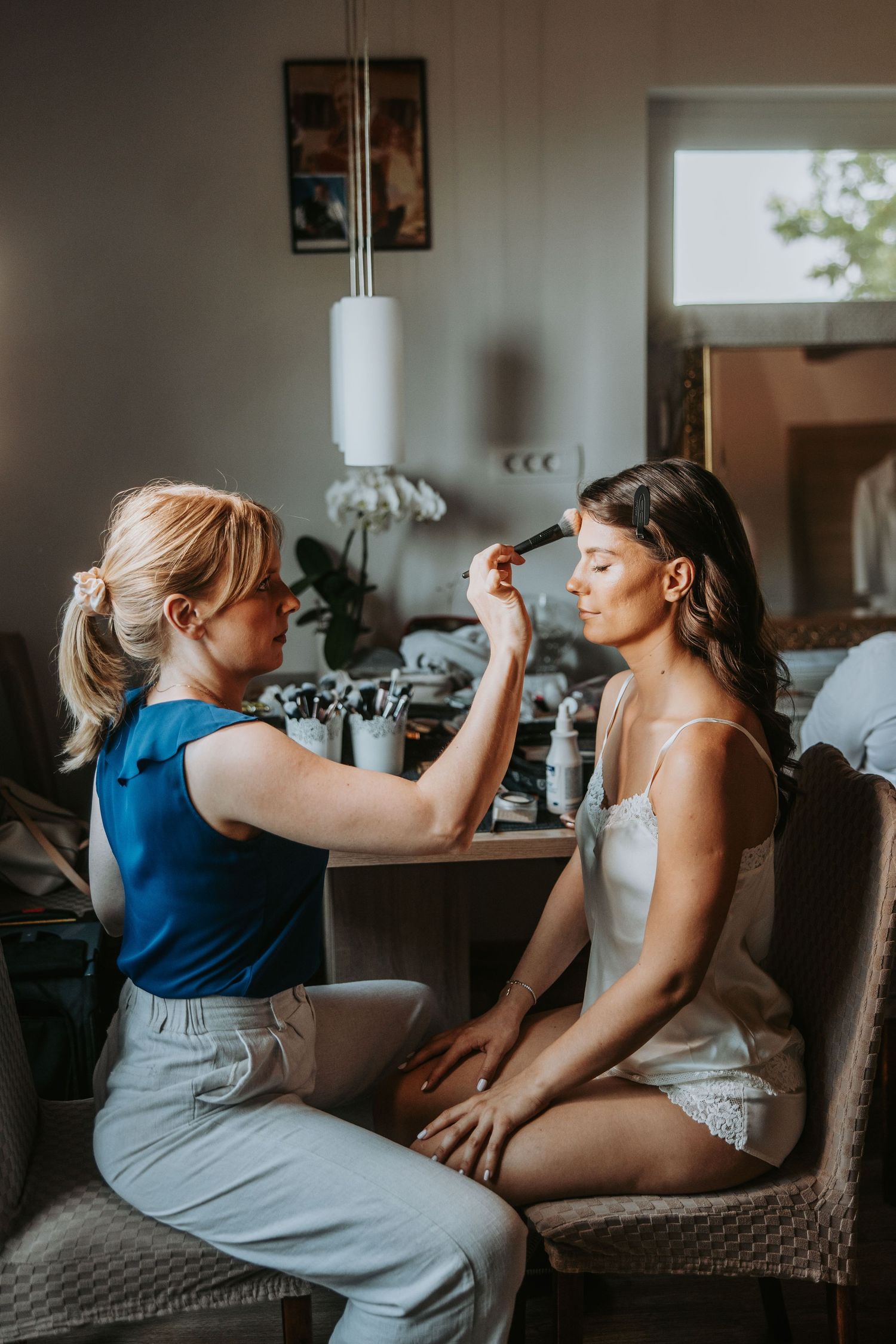 Makeup artist applying eyeshadow to a bride in white lingerie during wedding preparations indoors.