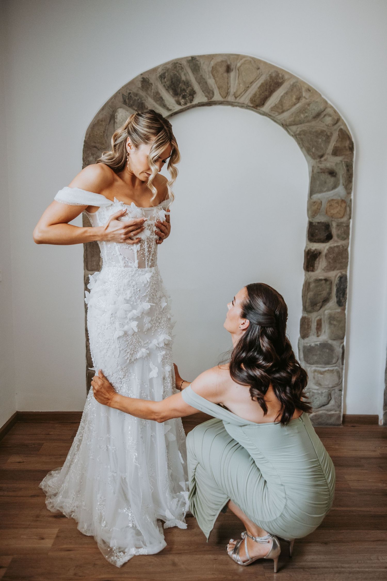 Bridesmaid in sage green dress helping bride into fitted lace wedding gown near a stone arch doorway.