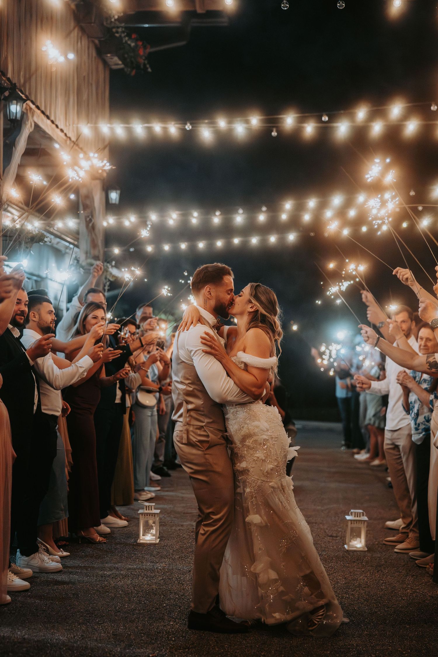 Bride and groom share a romantic kiss during a sparkler send-off under string lights at night.