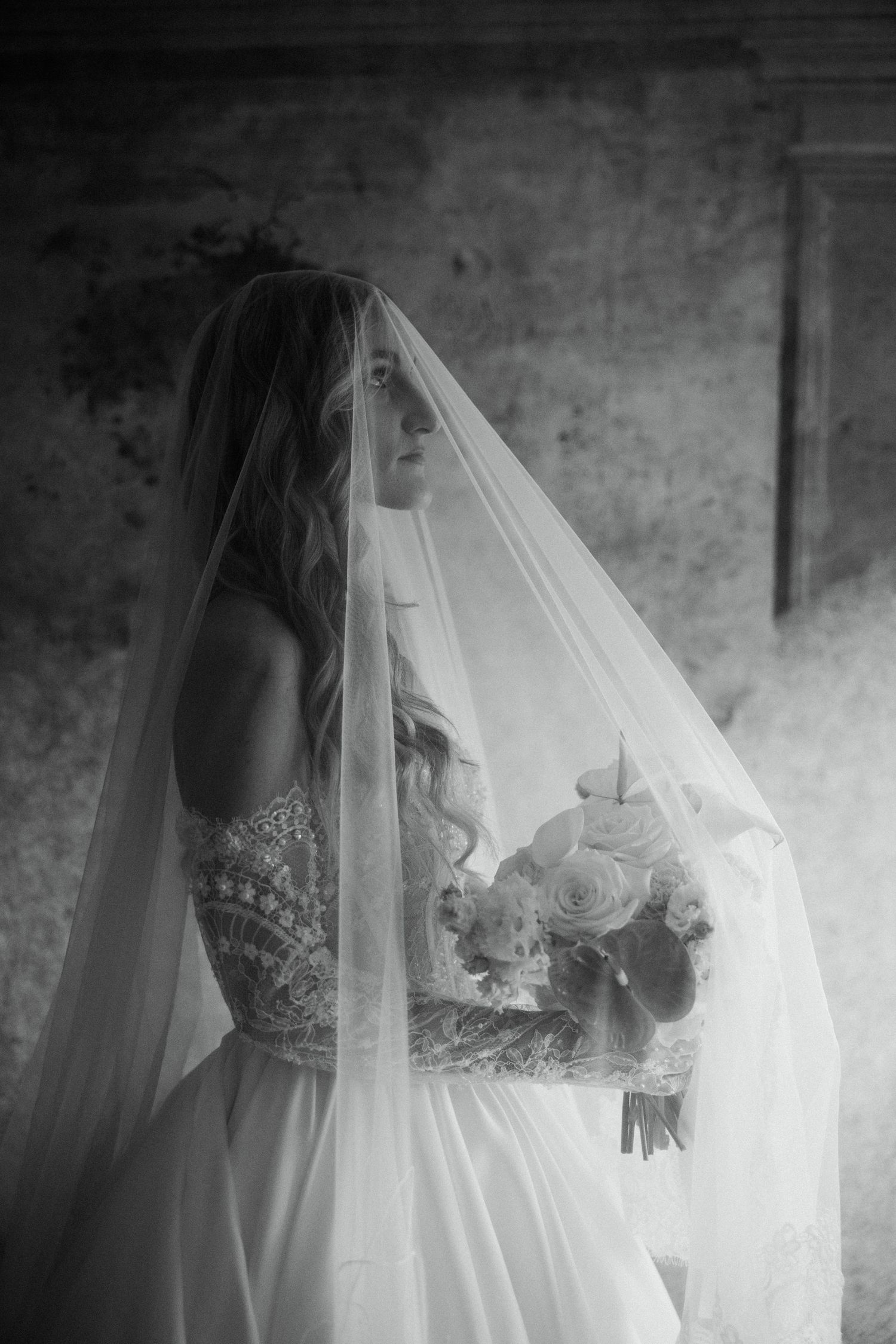 Romantic black and white portrait of bride in lace gown holding bouquet beneath flowing cathedral veil.