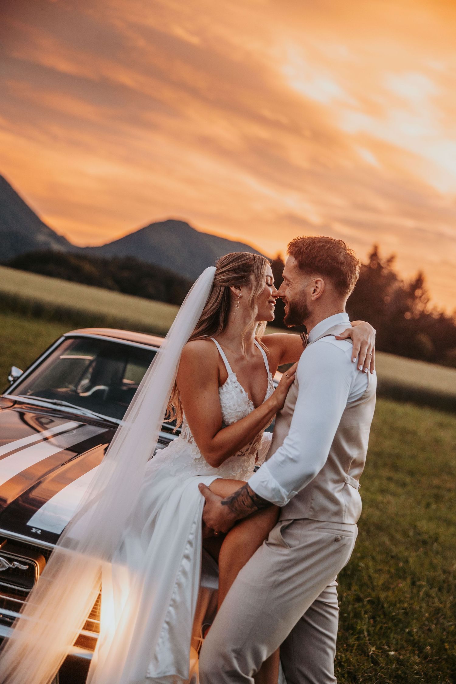Bride and groom share intimate embrace beside vintage Mustang with stunning orange sunset backdrop.