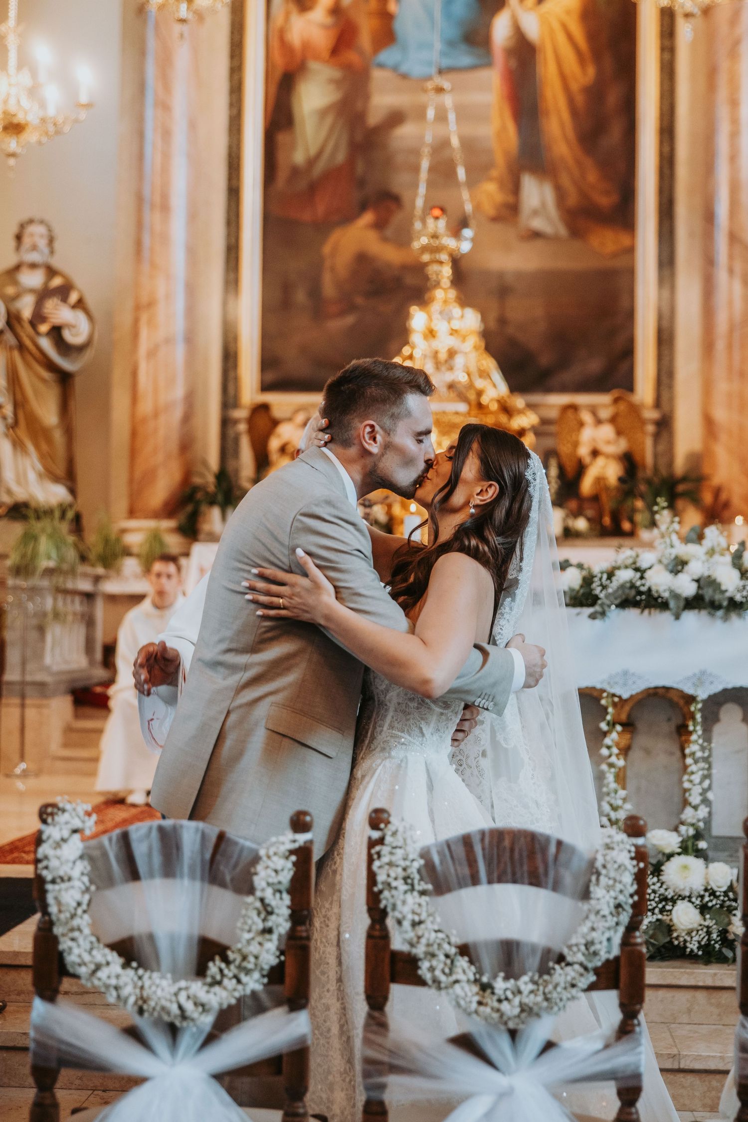 Newlyweds share first kiss at decorated church altar with golden religious artwork backdrop.