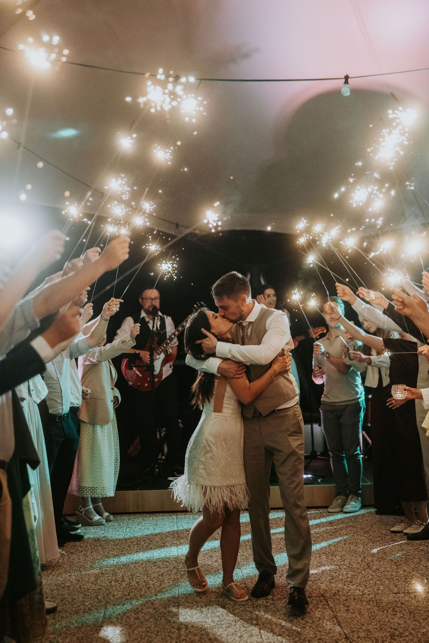 Bride and groom kiss under sparklers held by cheering wedding guests at reception.