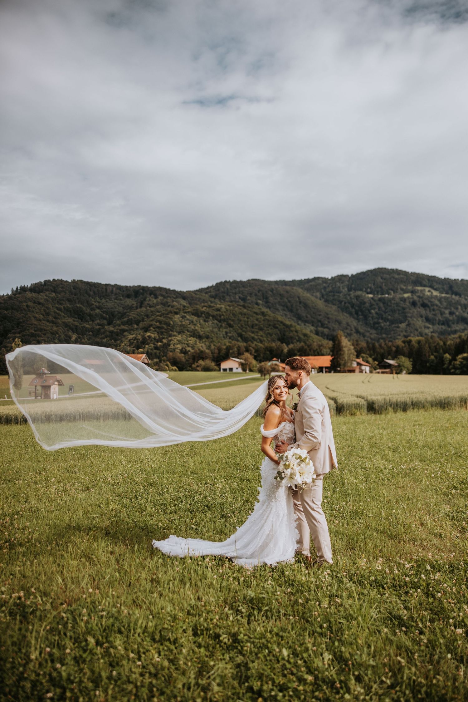 Bride and groom embrace in green meadow, veil flowing in wind, mountain backdrop on cloudy wedding day.