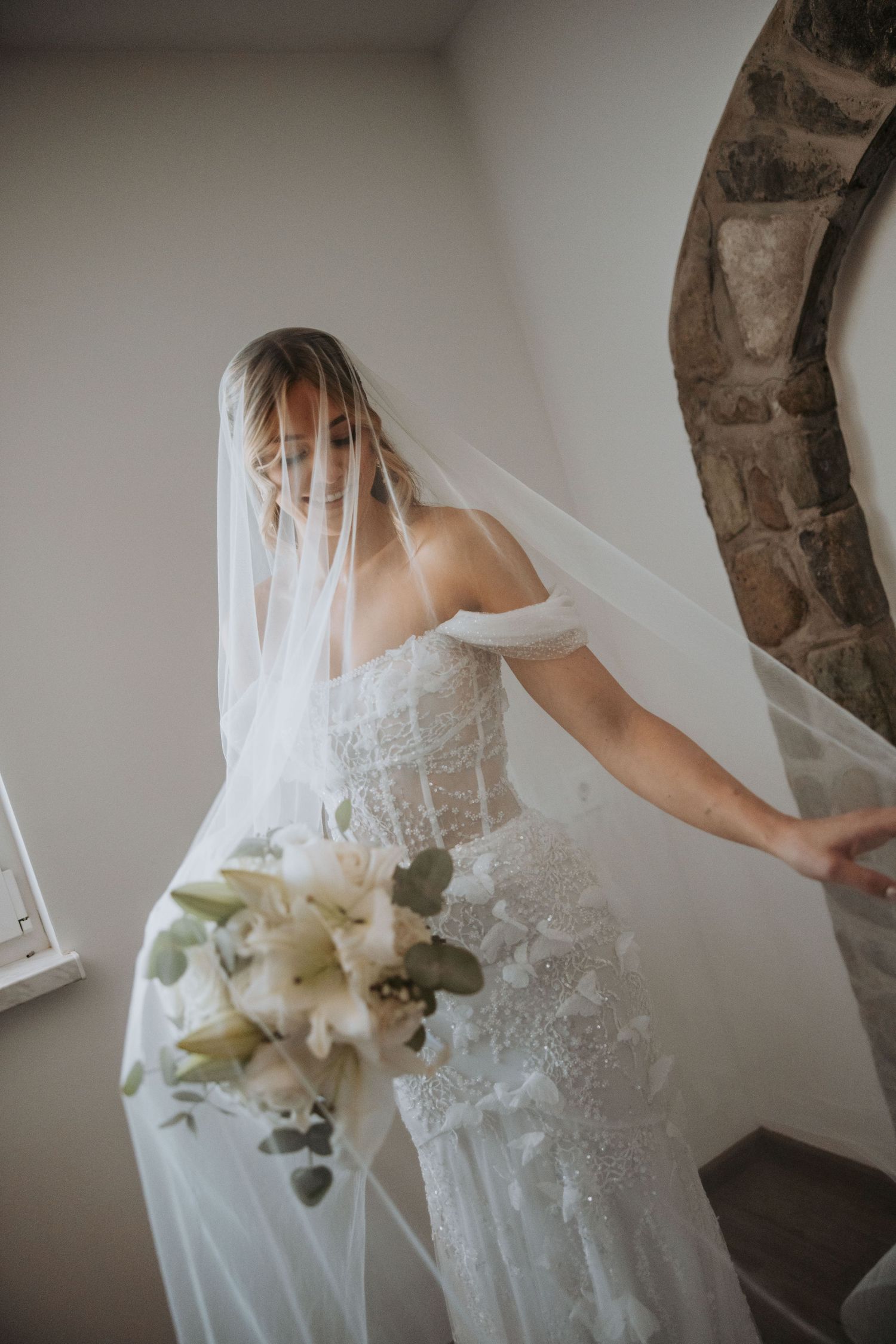 Bride in off-shoulder lace wedding dress holds white floral bouquet while adjusting flowing veil.
