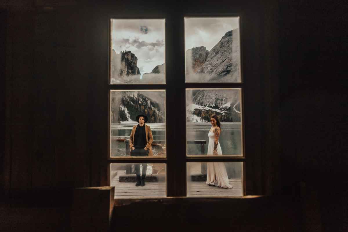 Bride and groom stand apart viewed through a dark window with mountain lake artwork panels.
