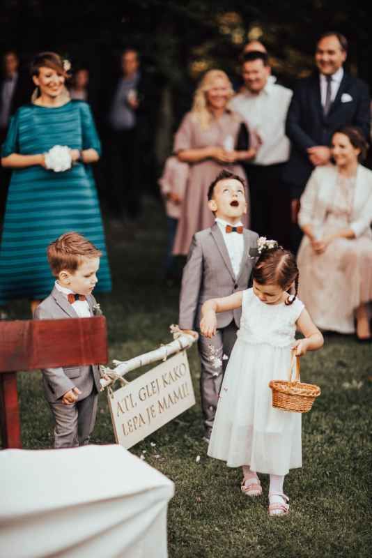 Young ring bearers and flower girl walking down wedding aisle holding decorative sign at outdoor ceremony.