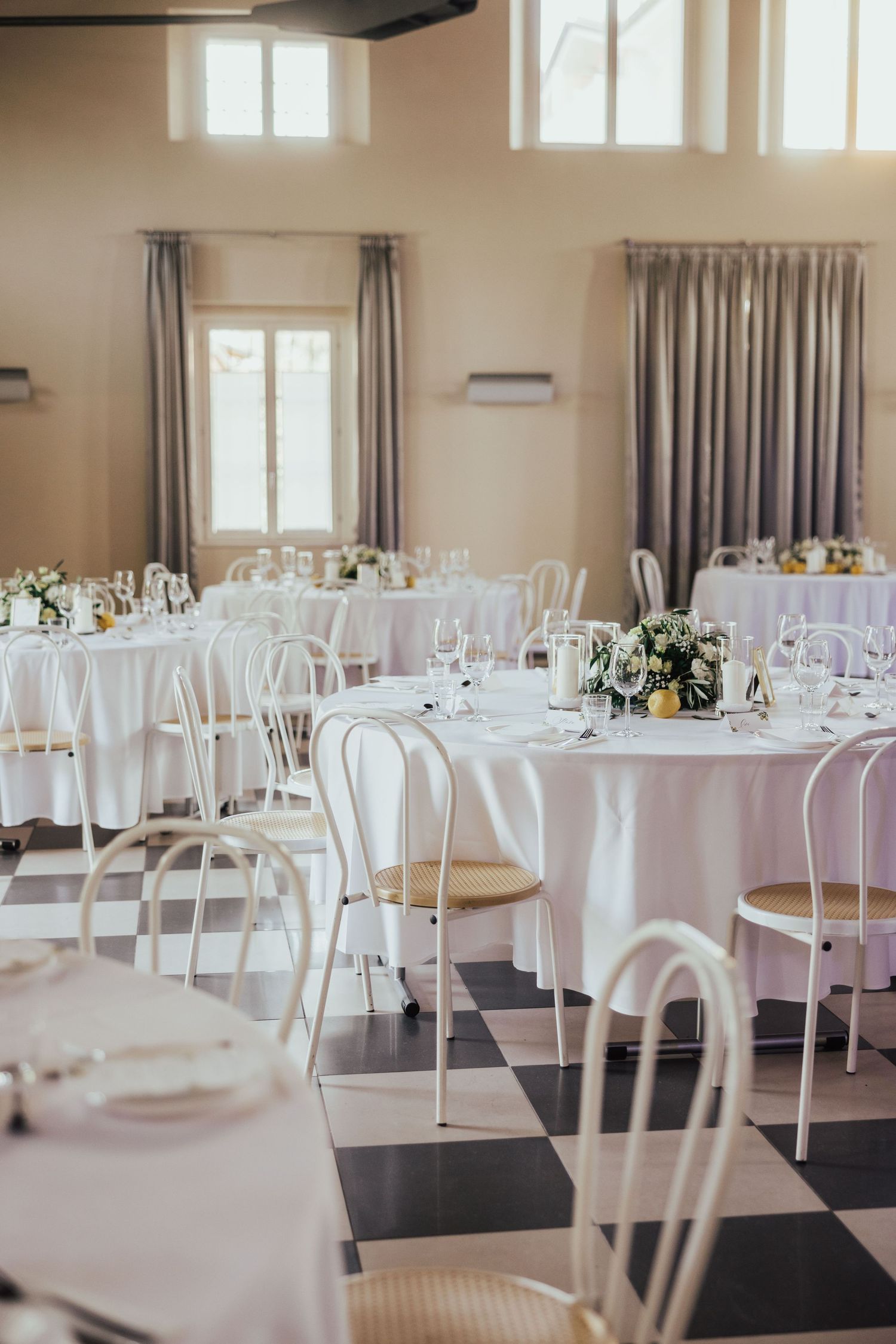 Elegant wedding reception hall with white round tables, bentwood chairs, floral centerpieces, and a classic black and white checkered floor.