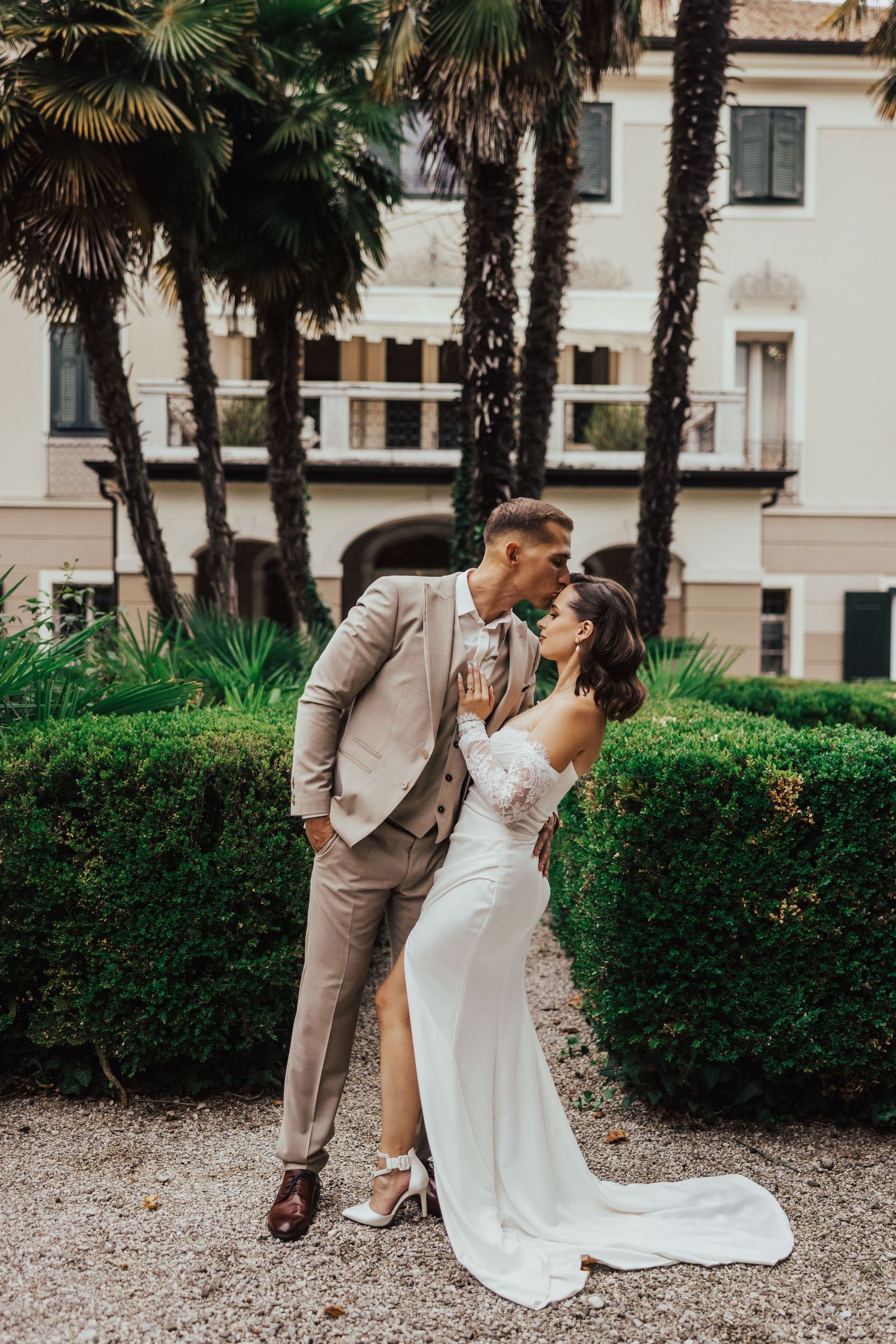 Bride and groom kissing in elegant garden with palm trees and villa backdrop on their wedding day.