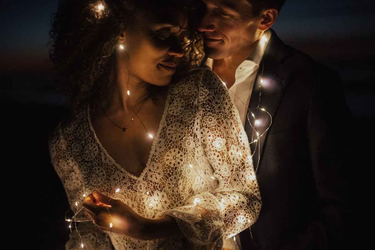 Romantic couple wrapped in glowing fairy lights at night, sharing an intimate moment in elegant wedding attire.