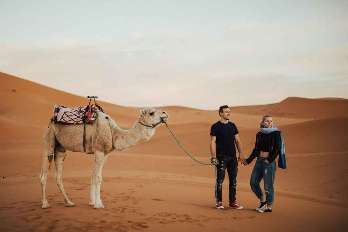 Couple holding hands with a saddled camel in the Sahara Desert, surrounded by golden sand dunes at sunset.