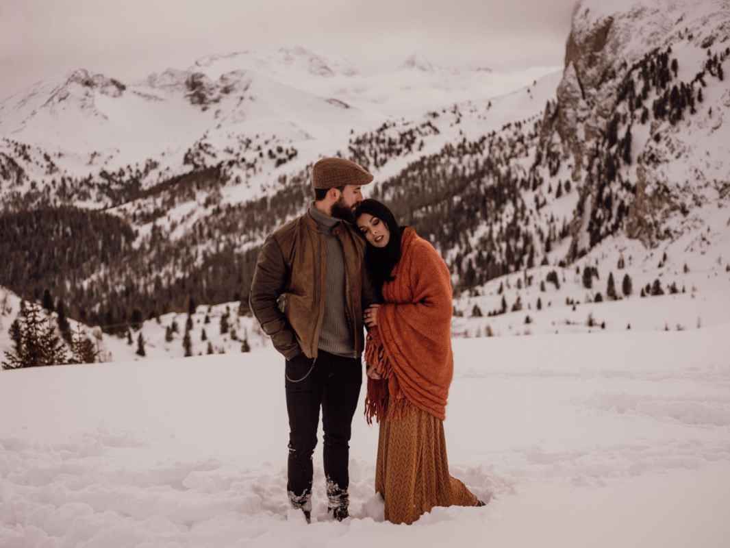 Couple embracing in snowy mountain landscape, woman wrapped in rust orange blanket during winter elopement.