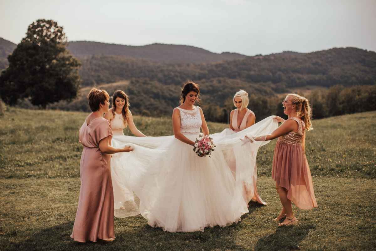 Bride in white gown laughs joyfully as four bridesmaids in pink dresses help arrange her train outdoors.