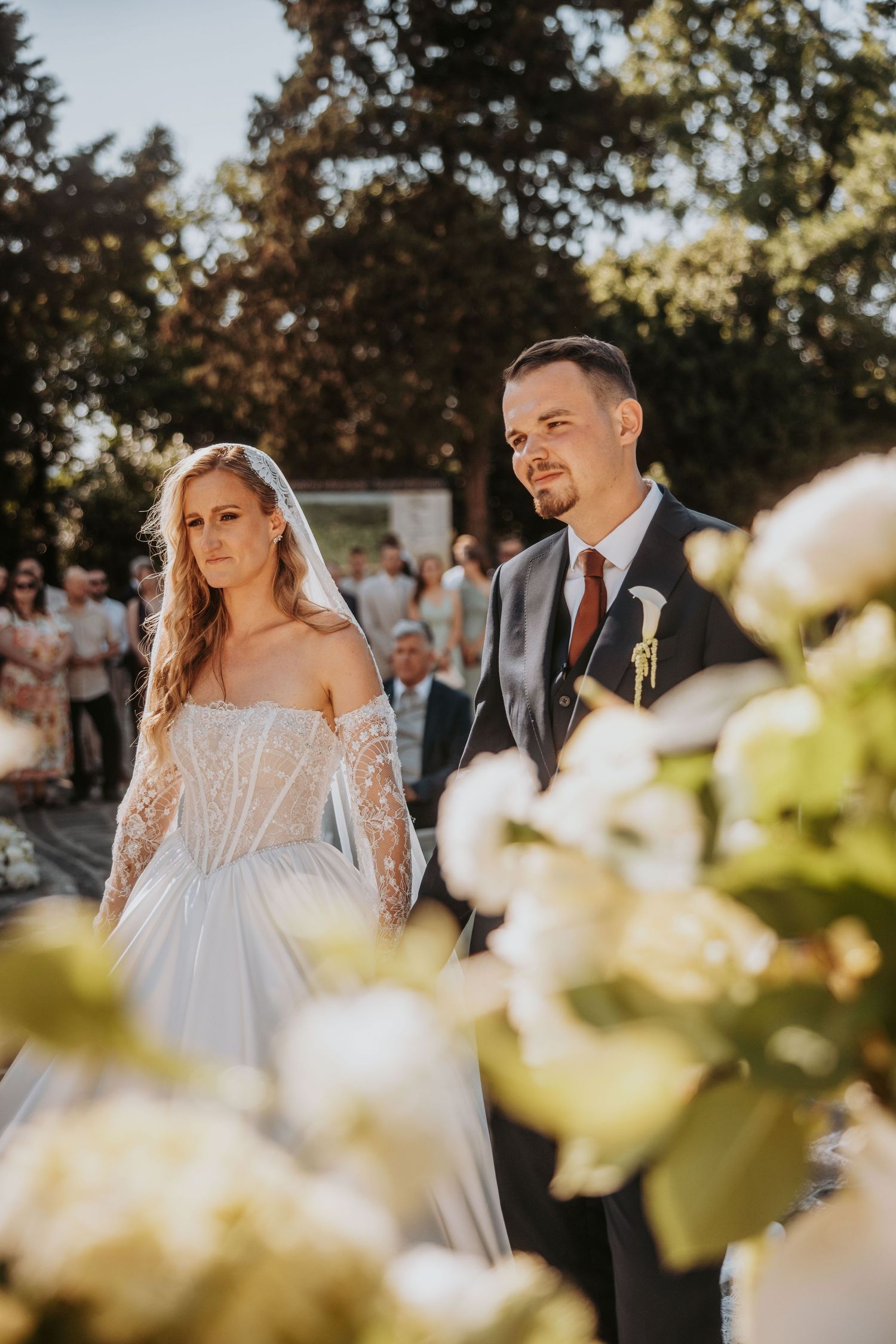 Bride and groom stand together at outdoor wedding ceremony surrounded by white floral arrangements and guests.