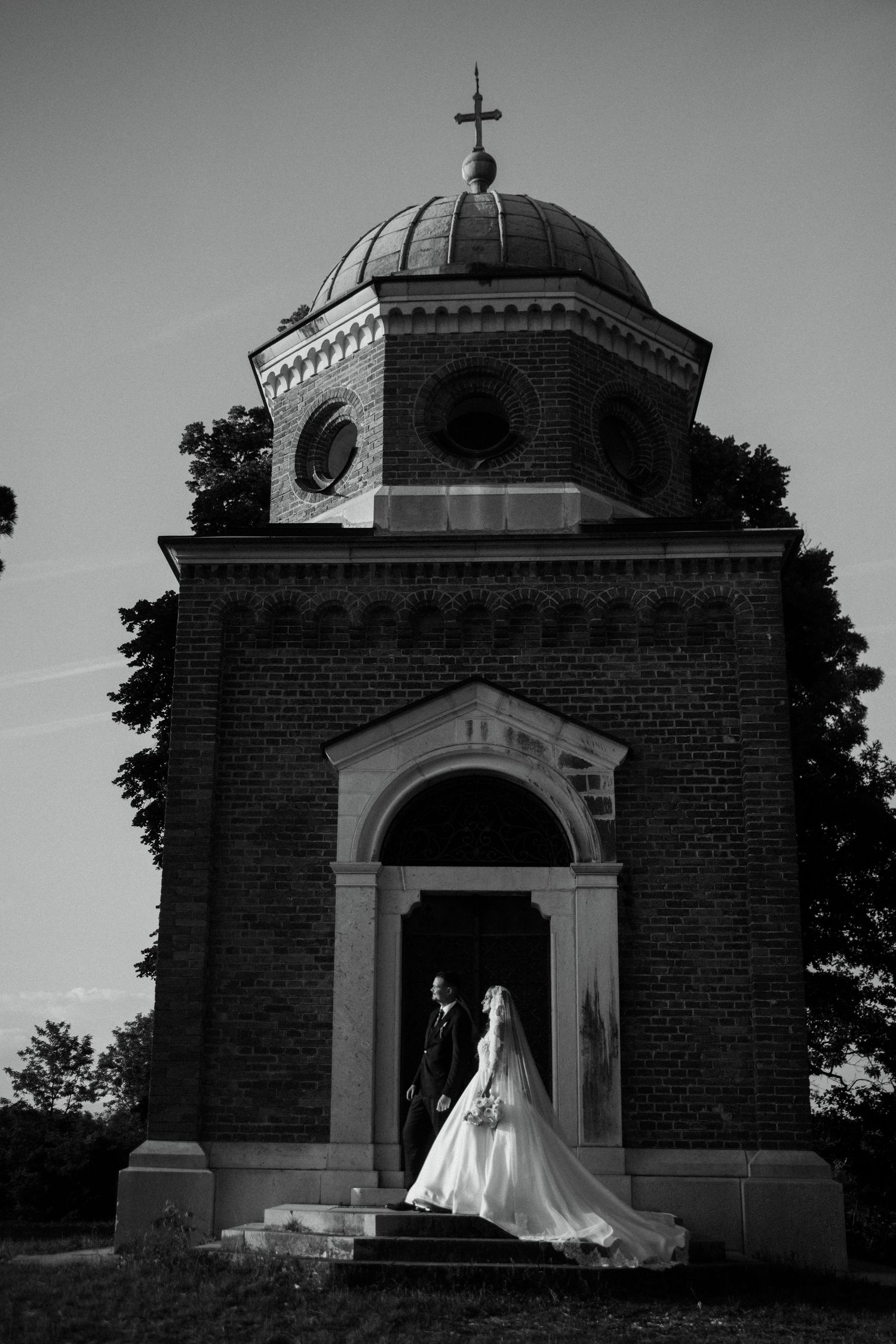 Black and white photo of bride and groom standing in arched doorway of historic brick chapel with domed roof.