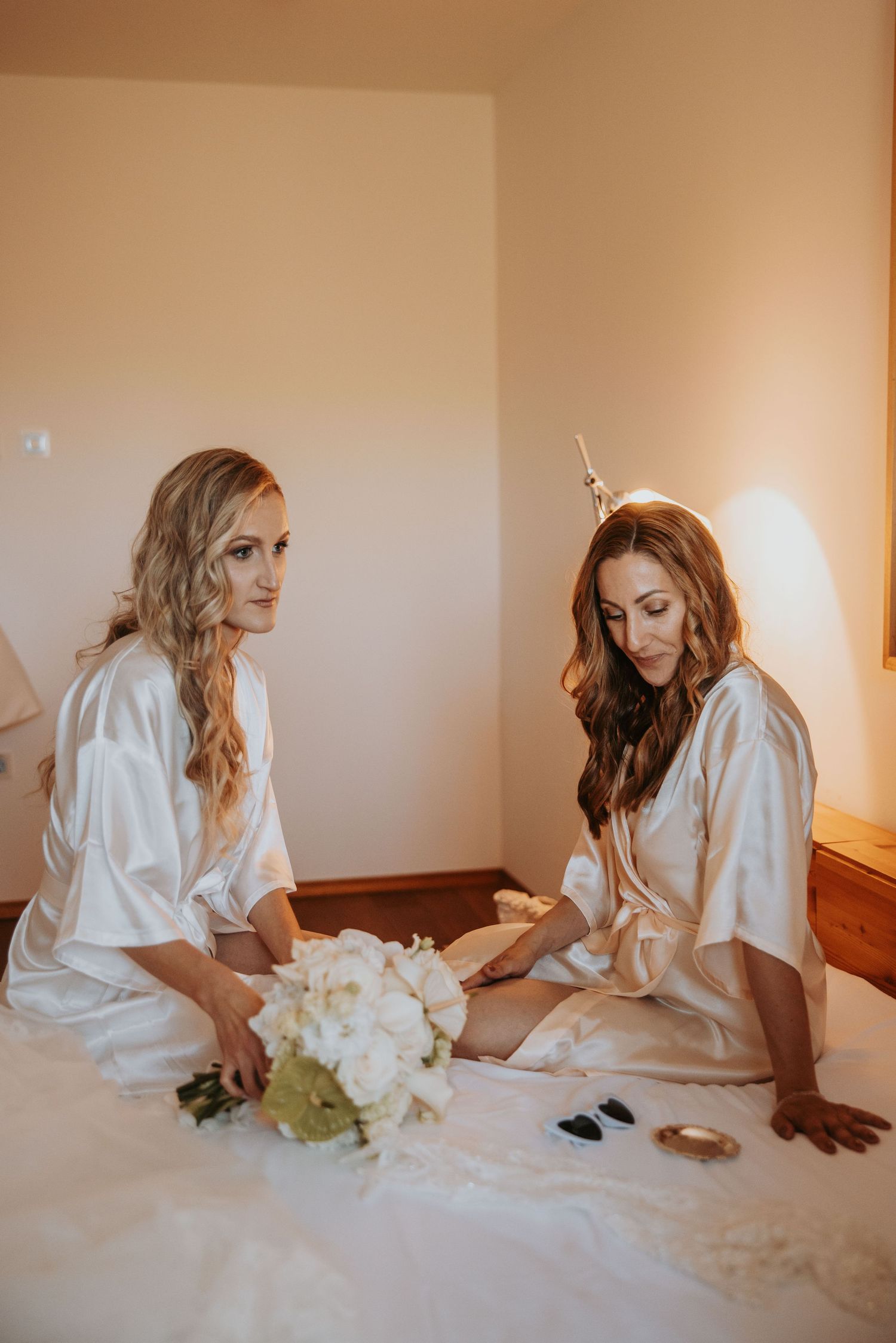 Two bridesmaids in white satin robes sit on a bed with a white bouquet during wedding preparations.