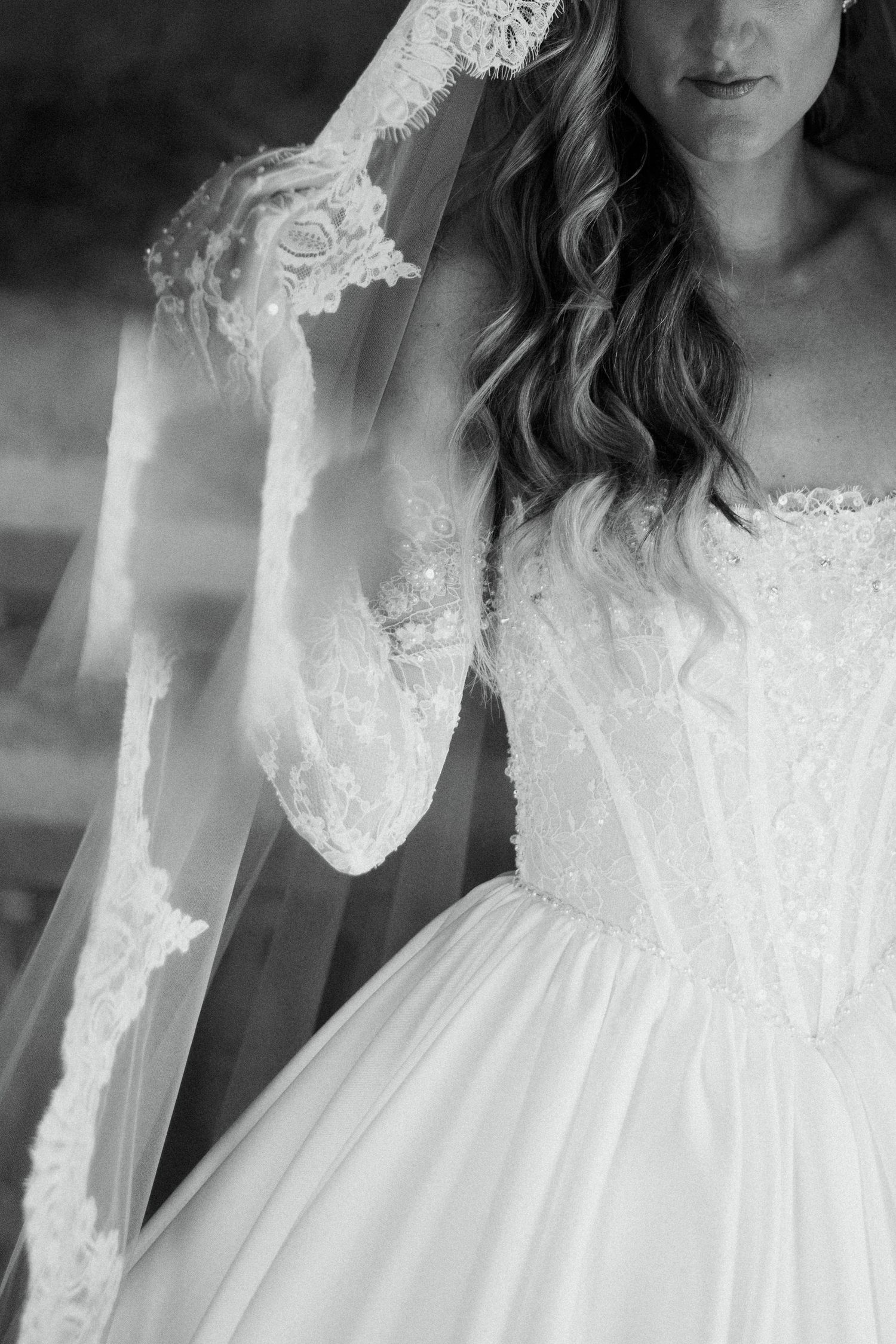 Black and white photo of a bride in a lace corset ballgown holding her lace-trimmed veil with long lace sleeves.