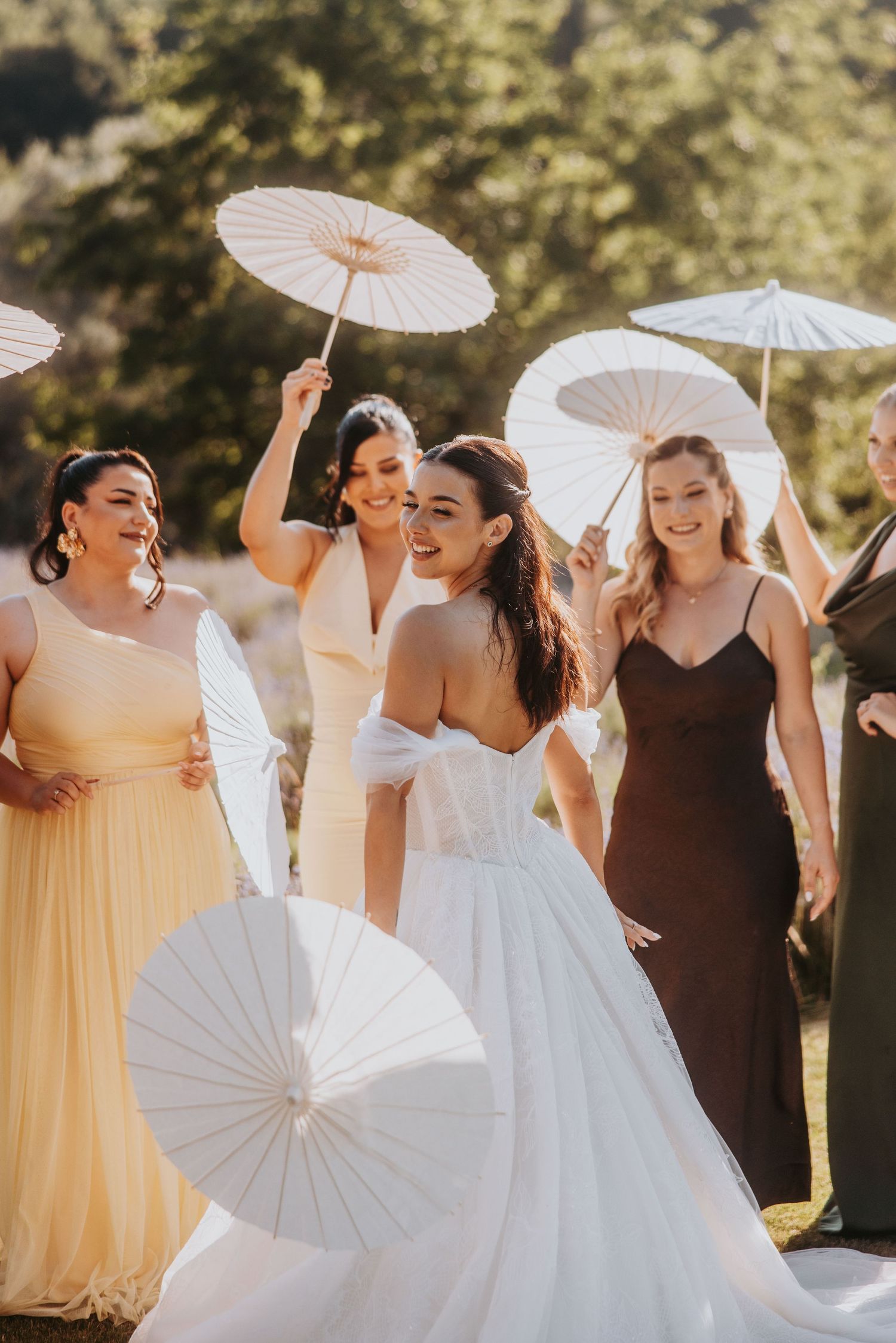 Bride in white gown twirling with bridesmaids holding paper parasols in golden sunlight at outdoor wedding.