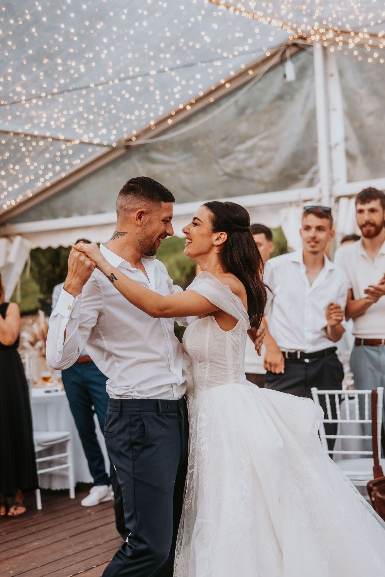 Smiling bride and groom share their first dance under twinkling fairy lights at an outdoor wedding reception.