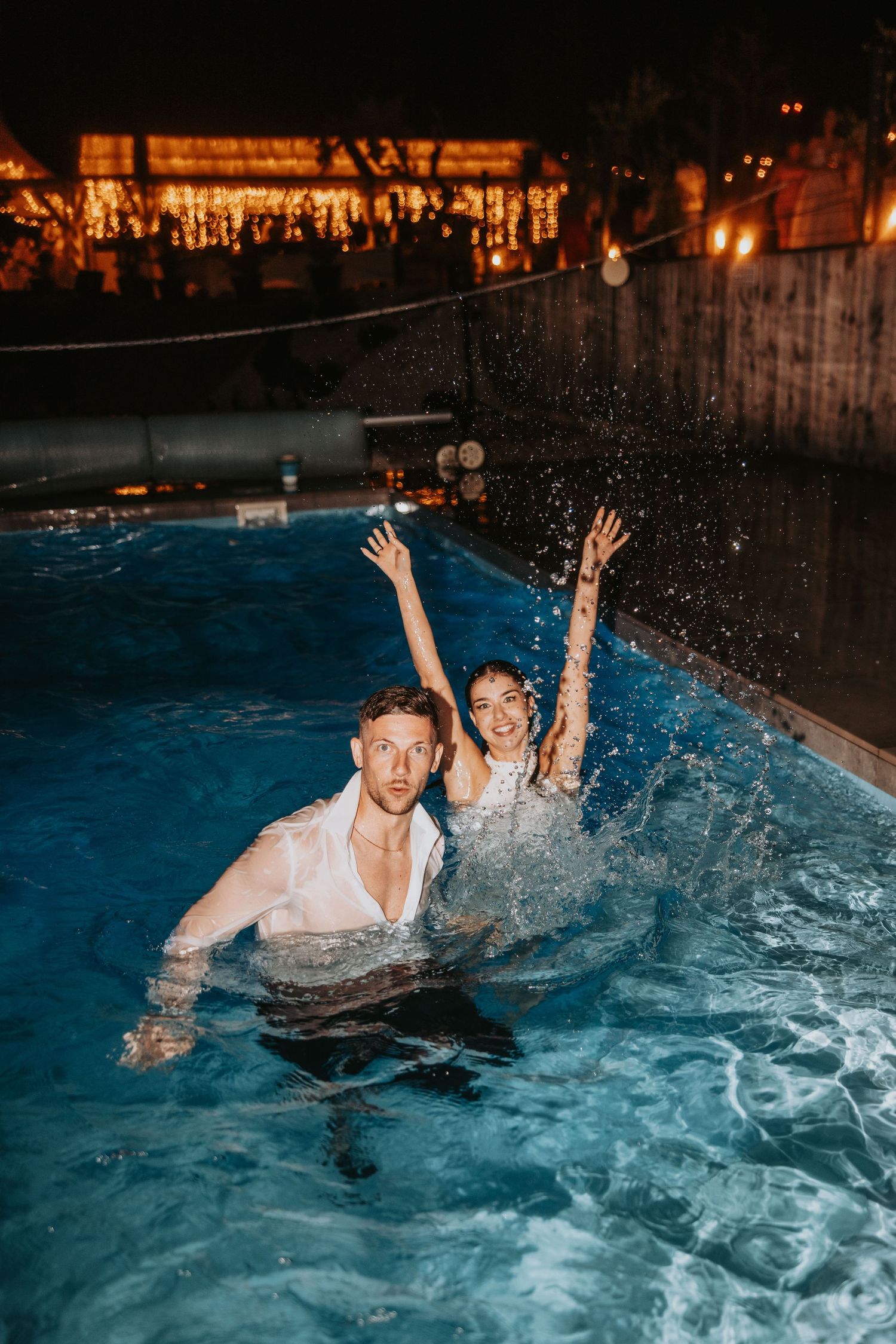 Newlyweds splash joyfully in illuminated pool at night still wearing wedding attire.