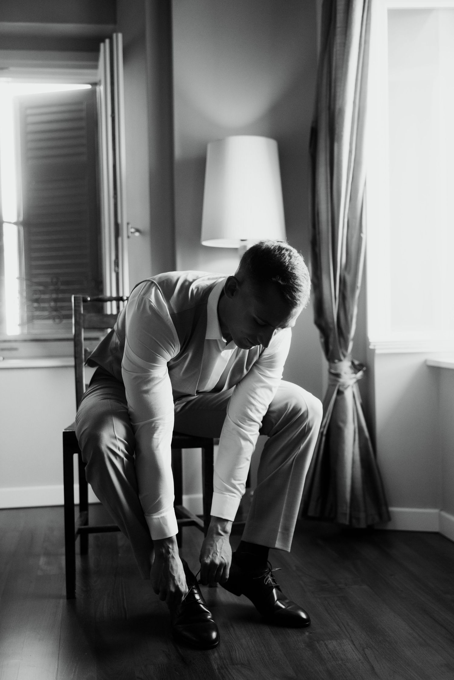 Black and white photo of groom sitting in chair lacing up dress shoes before wedding ceremony.
