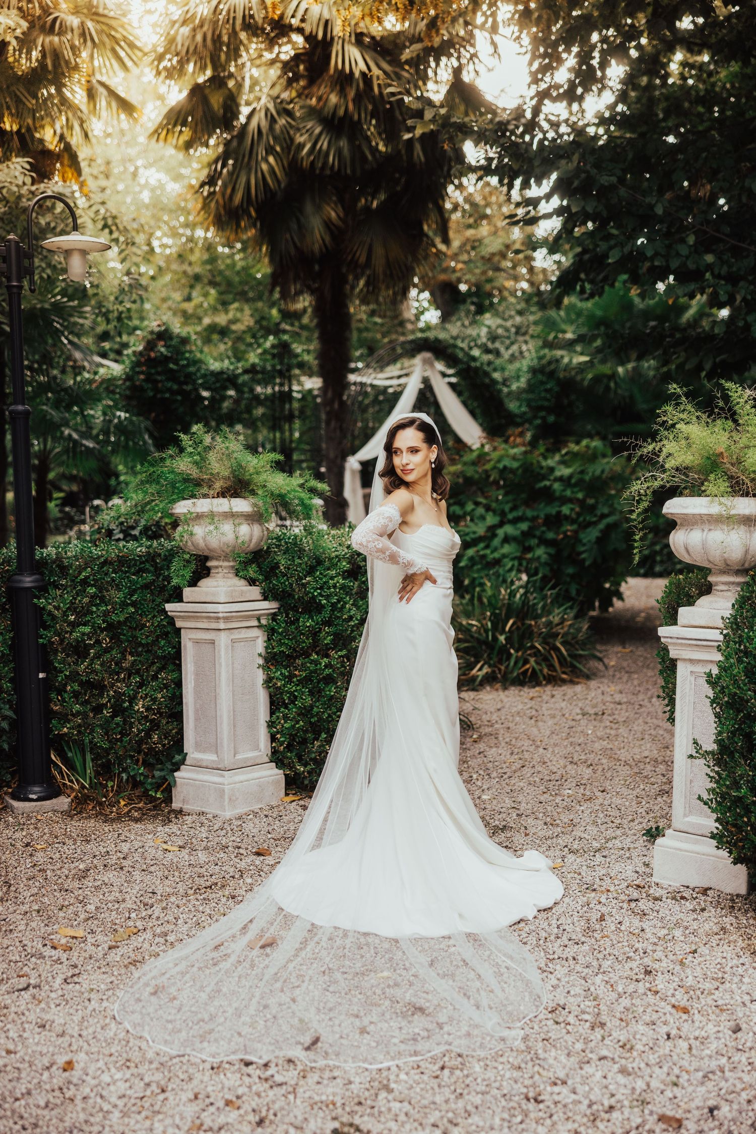 Bride in a fitted white gown and long veil poses gracefully on a gravel path in a lush garden setting.