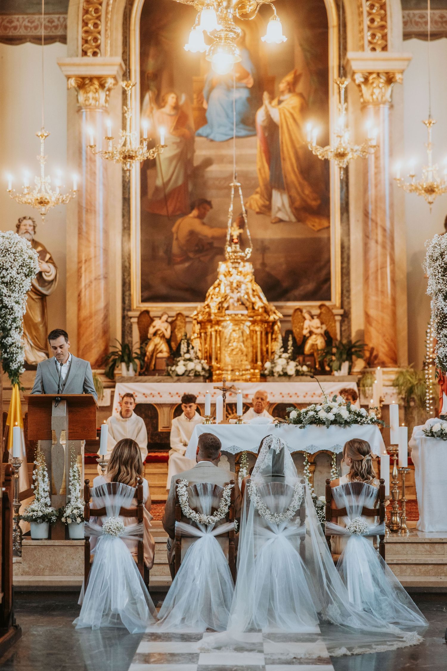 Elegant church wedding ceremony with bridesmaids in white gowns seated before ornate golden altar with floral arrangements.