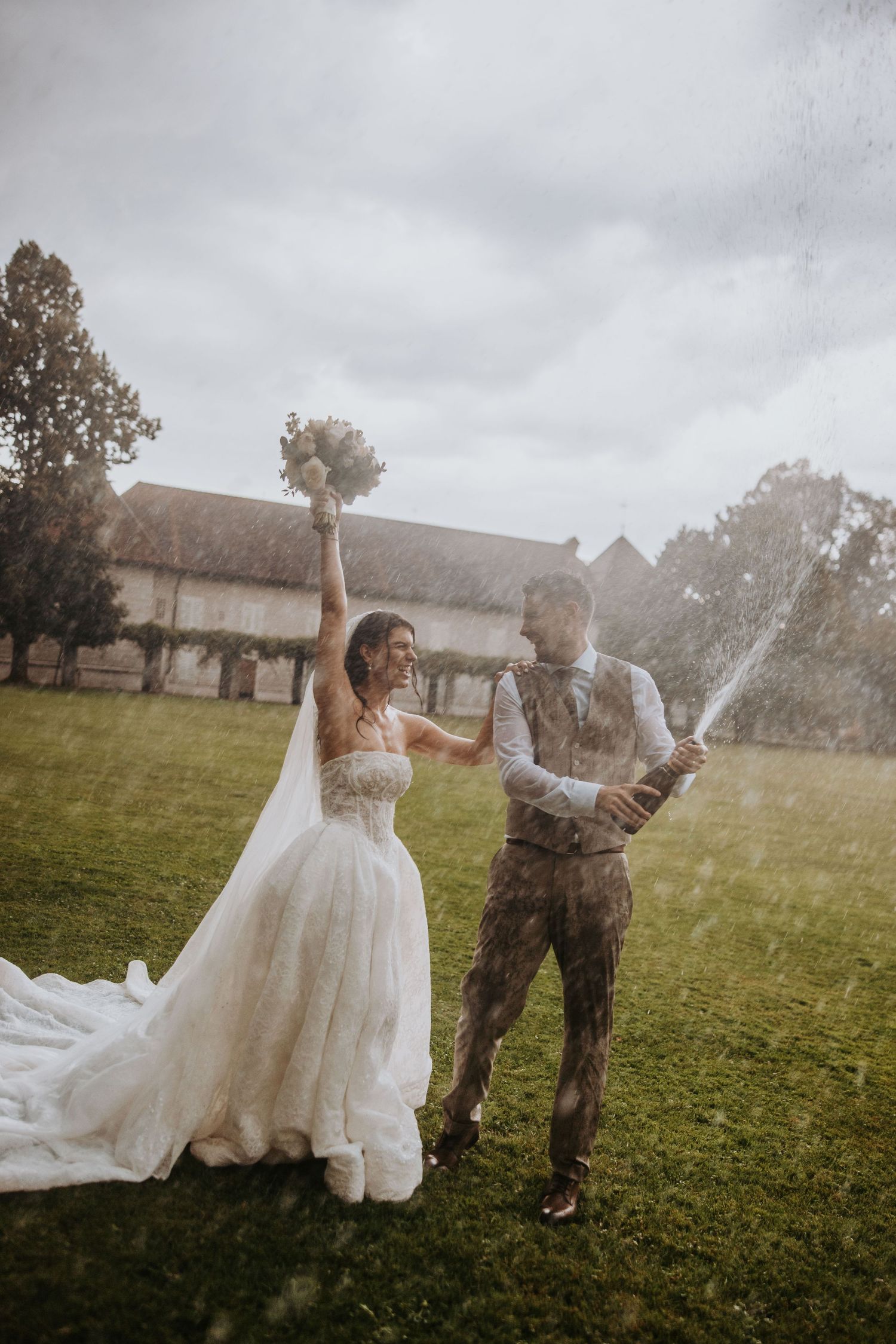 Joyful bride and groom celebrating outdoors popping champagne bottle in a lush green garden on their wedding day.