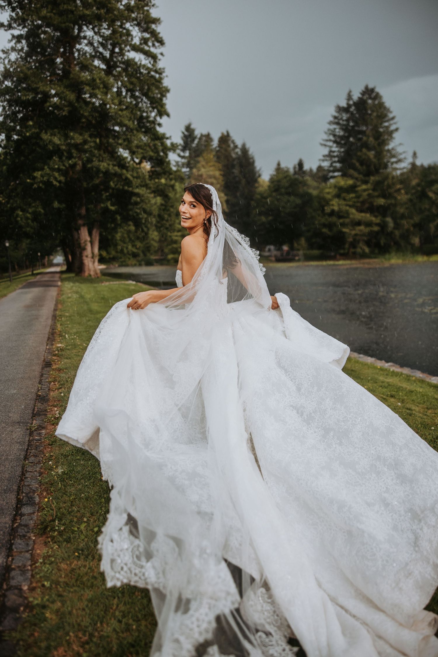 Bride in flowing lace gown and veil twirls near lake in dramatic outdoor portrait.