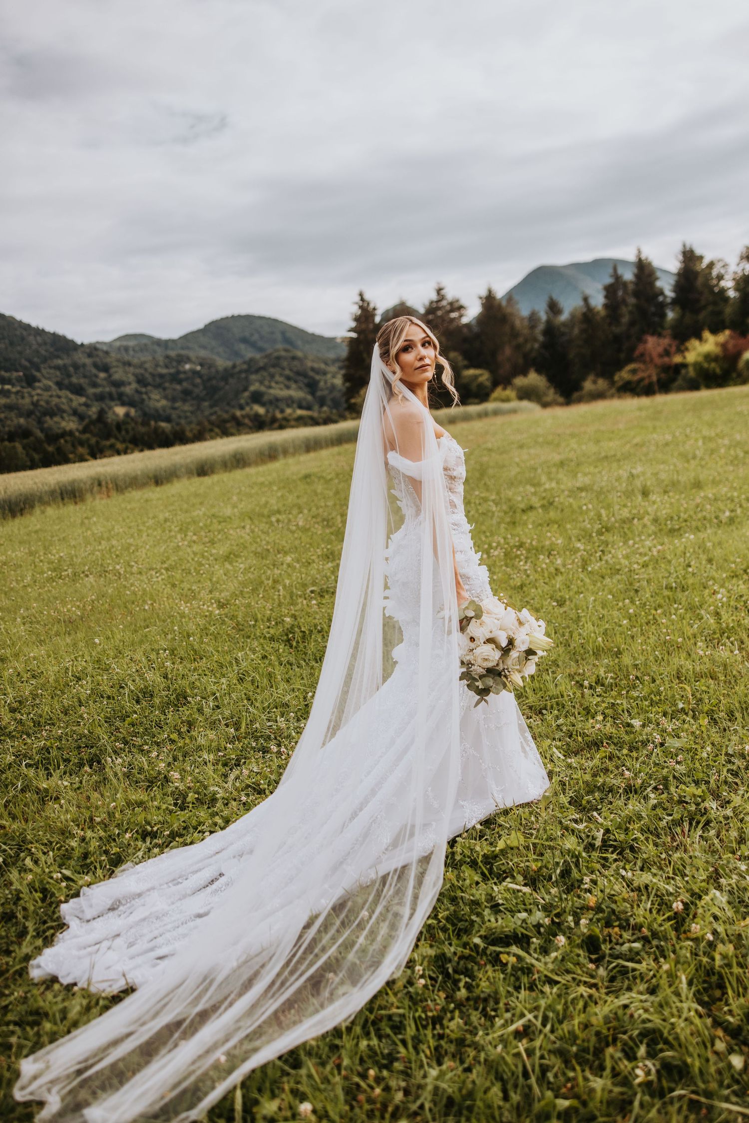 Bride in a flowing white lace gown and long veil stands in a green meadow with mountain backdrop.