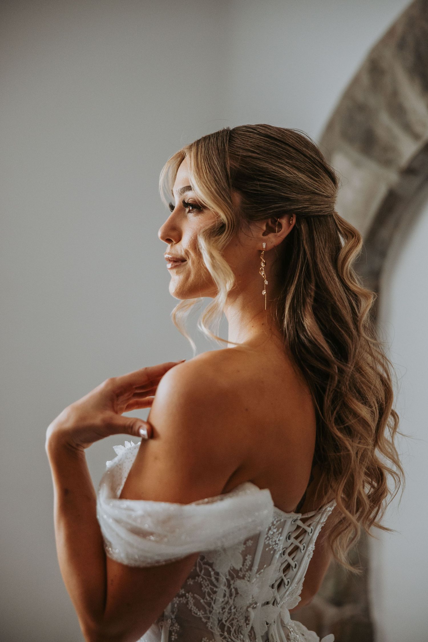 Elegant bride in off-shoulder lace wedding dress with wavy hair and drop earrings in soft light.