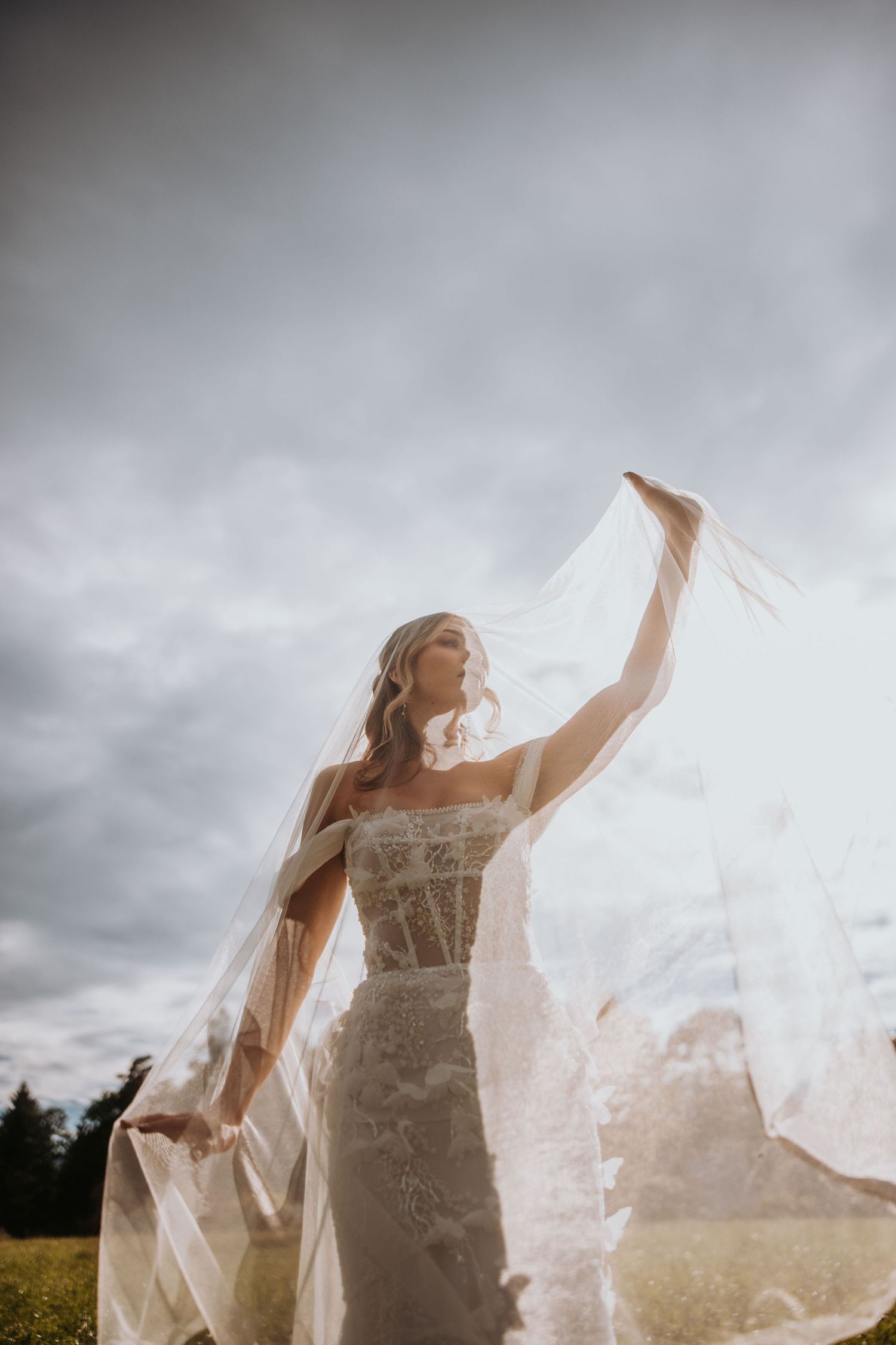 Bride in floral lace gown tosses veil in wind under dramatic cloudy sky in outdoor wedding portrait.