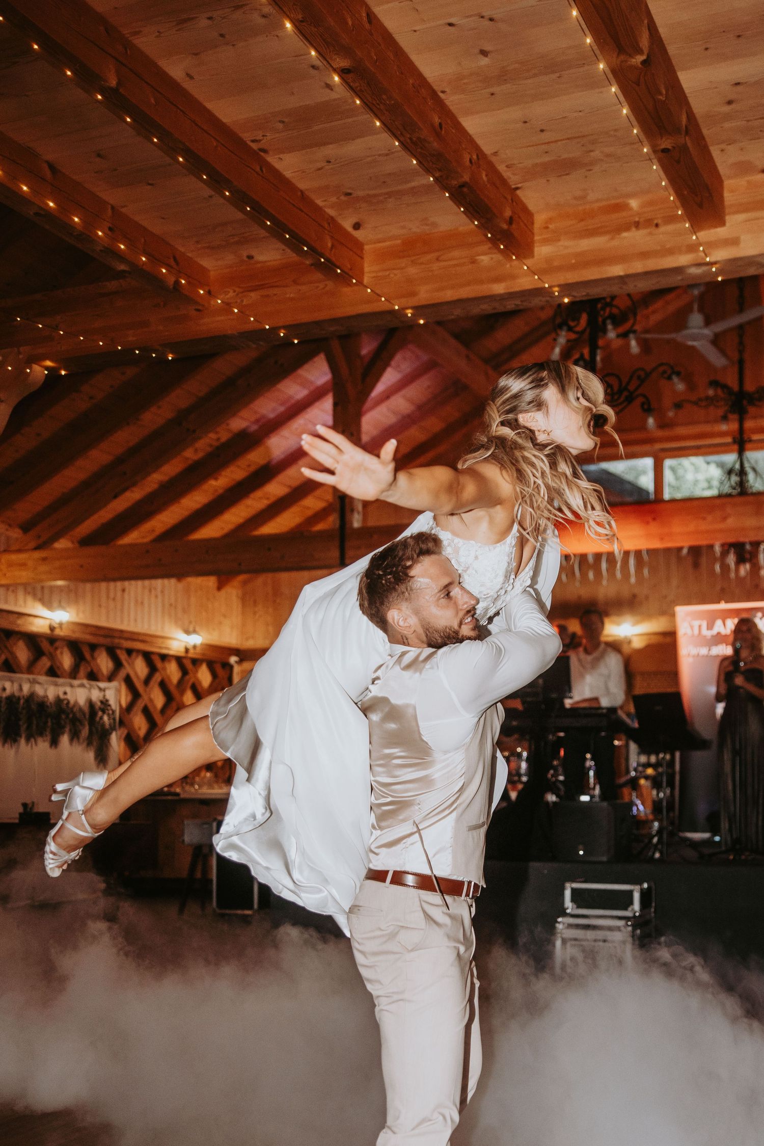 Groom lifts bride dramatically on smoky dance floor during energetic first dance at rustic wedding reception.