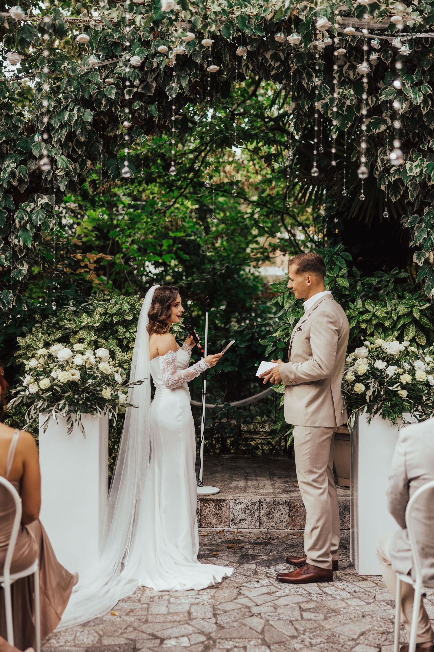 Bride and groom exchange vows under floral arch with white roses in lush outdoor garden ceremony.