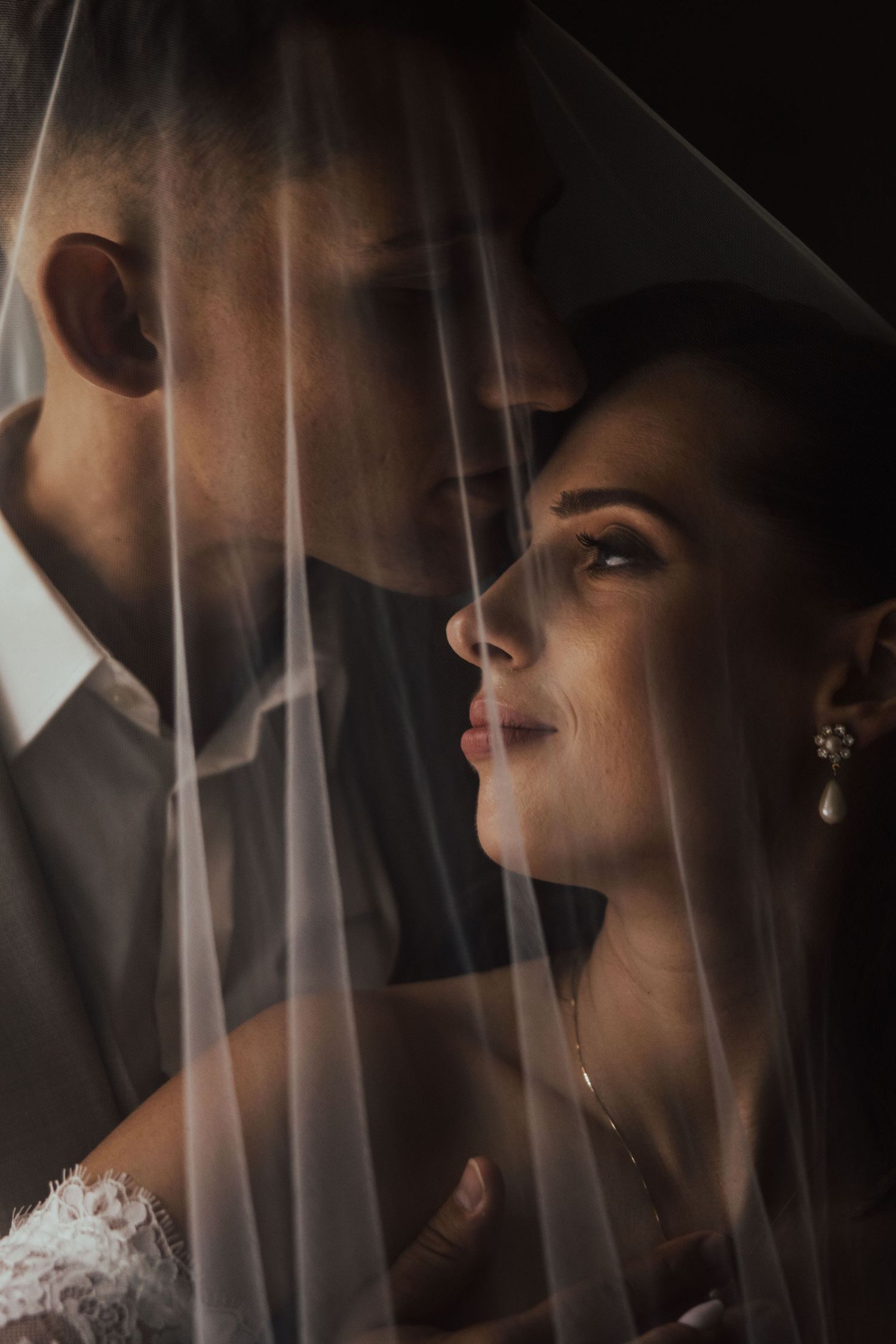 A bride and groom share an intimate moment under a flowing wedding veil in a romantic, softly lit portrait.