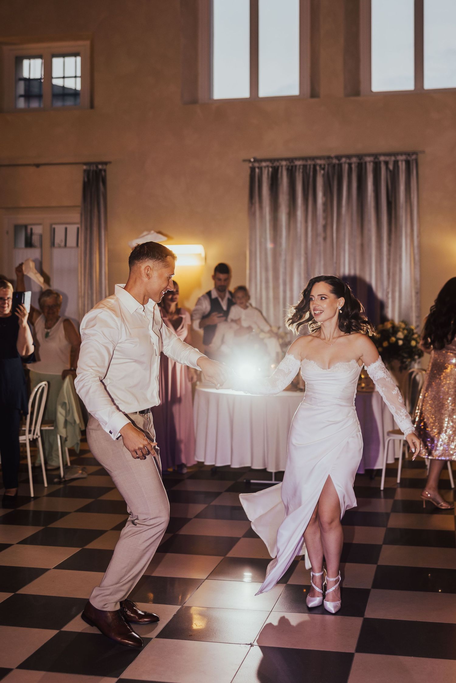 Bride and groom share a joyful first dance on a checkered floor surrounded by wedding guests in a warmly lit reception hall.