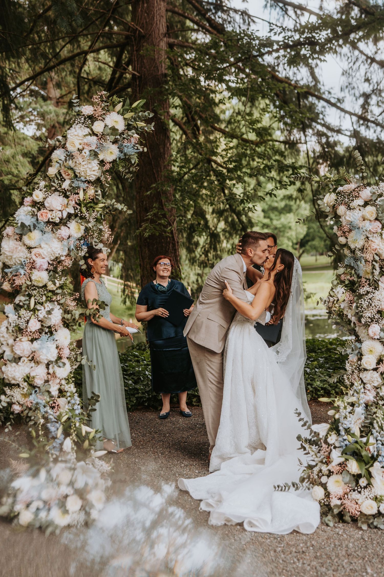 Bride and groom share first kiss under floral arch at outdoor wedding ceremony.