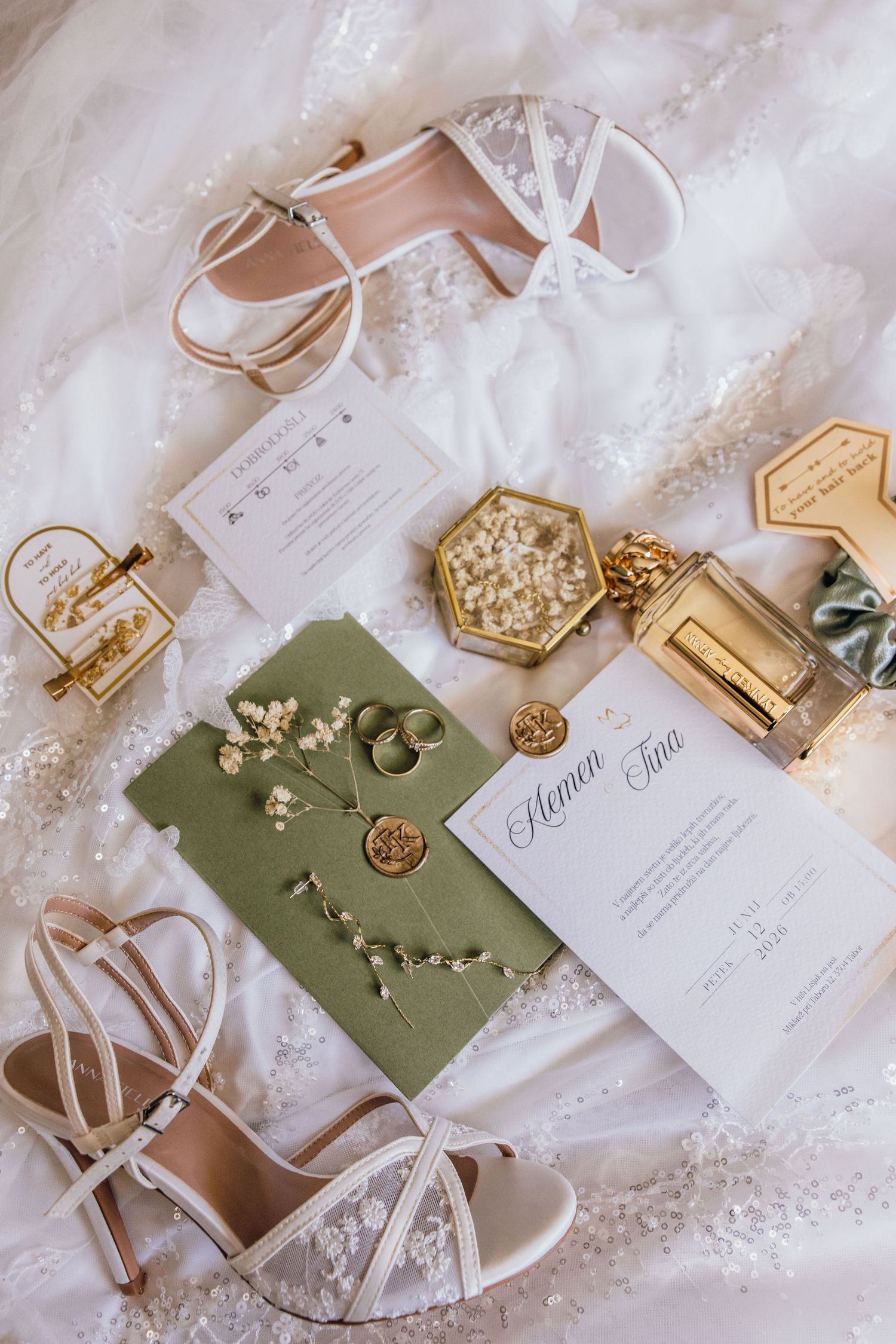 Elegant wedding flat lay with sage green invitation suite, gold rings, white heels, perfume bottle, and floral accents on lace fabric.