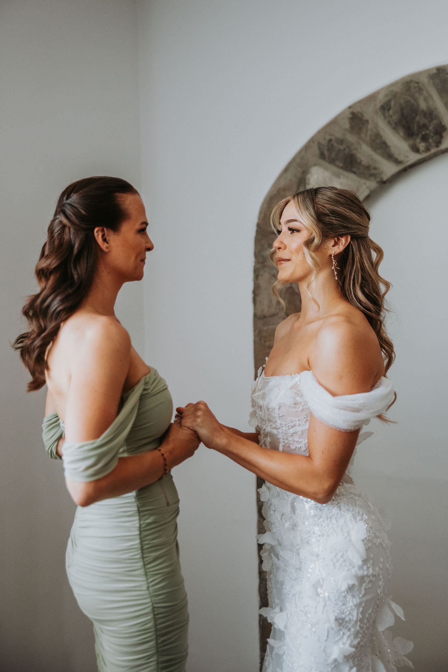 Bride in off-shoulder lace gown holds hands with bridesmaid in sage green dress near stone arch.