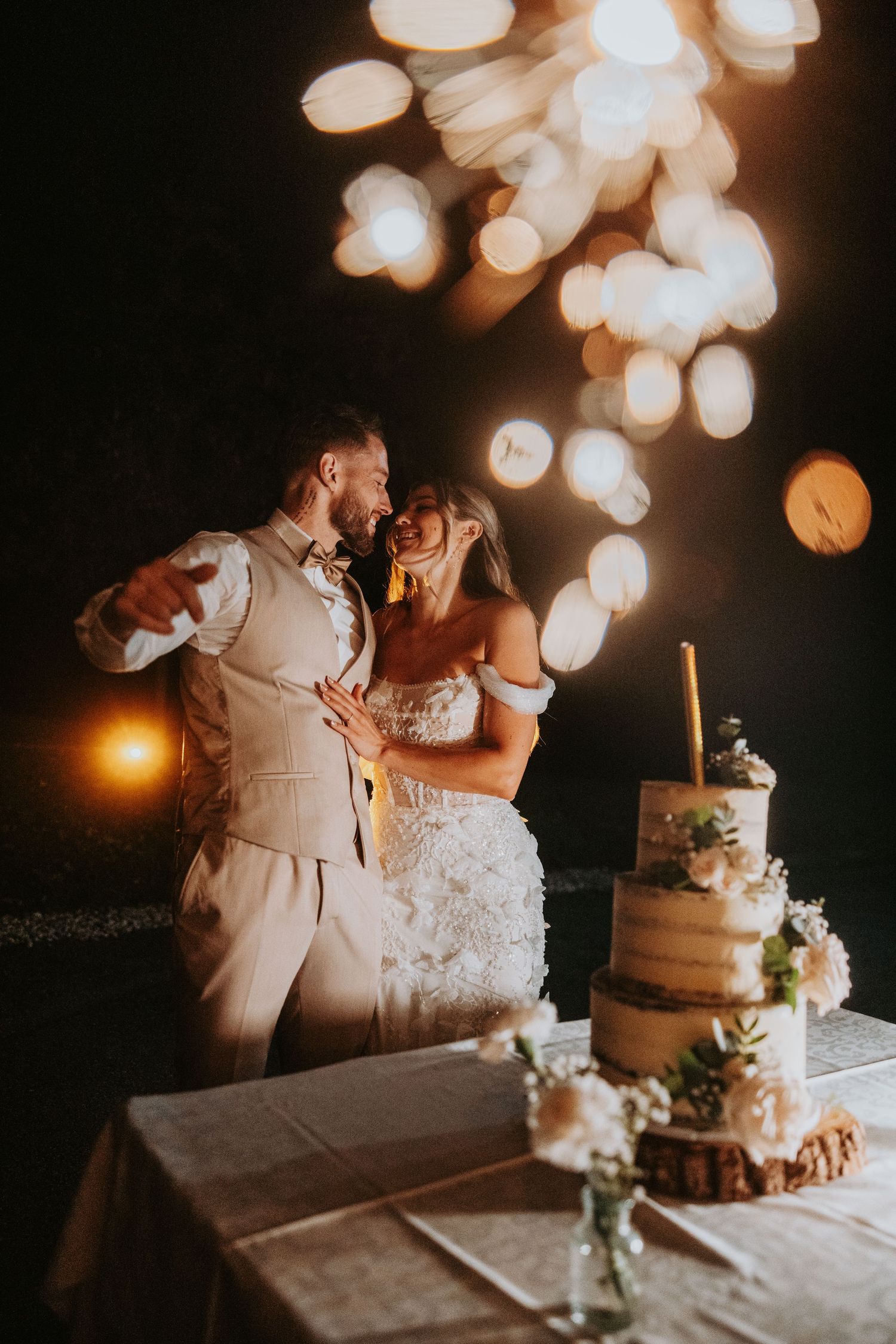 Bride and groom embrace near a three-tier wedding cake with bokeh lights glowing behind them.