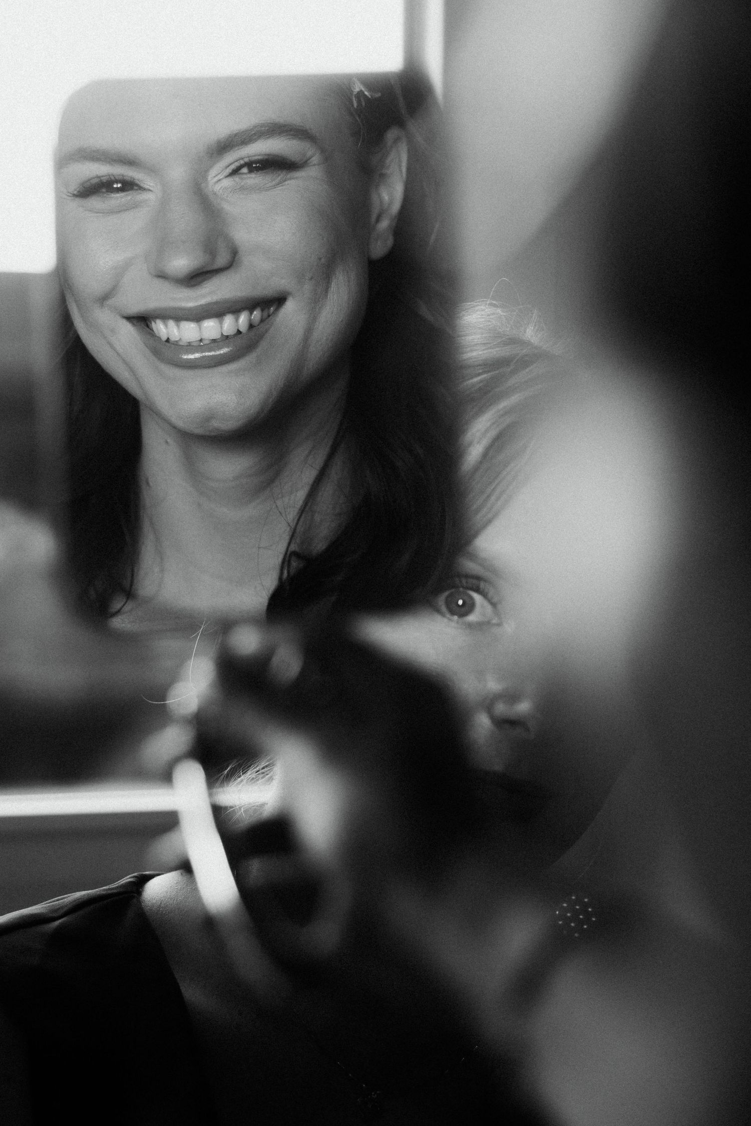 Black and white photo of a smiling woman with long hair holding a cigarette, with a child visible in the background.