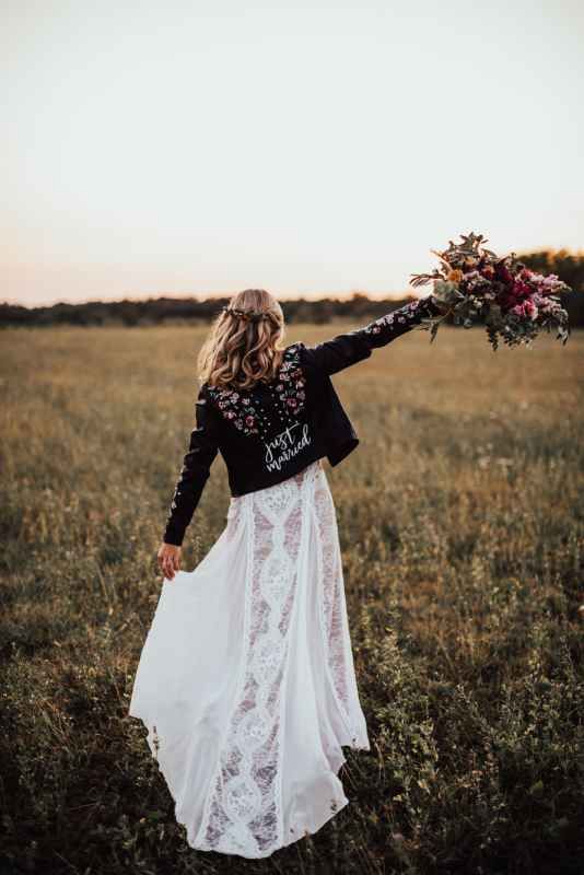 Bride in white lace gown and embroidered black leather jacket holding bouquet in golden field at sunset.