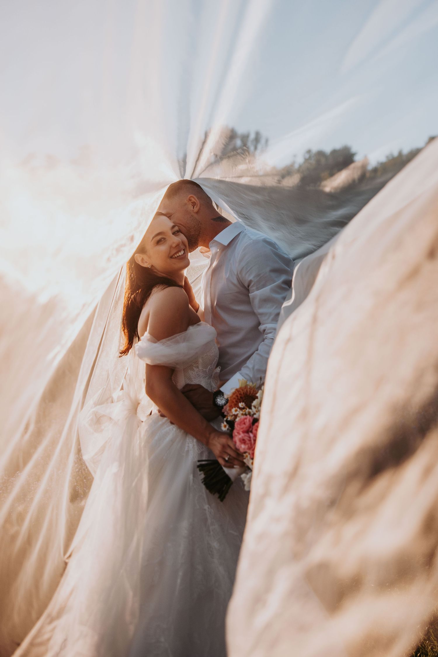 Bride and groom sharing a romantic moment under a billowing veil in warm golden sunlight.