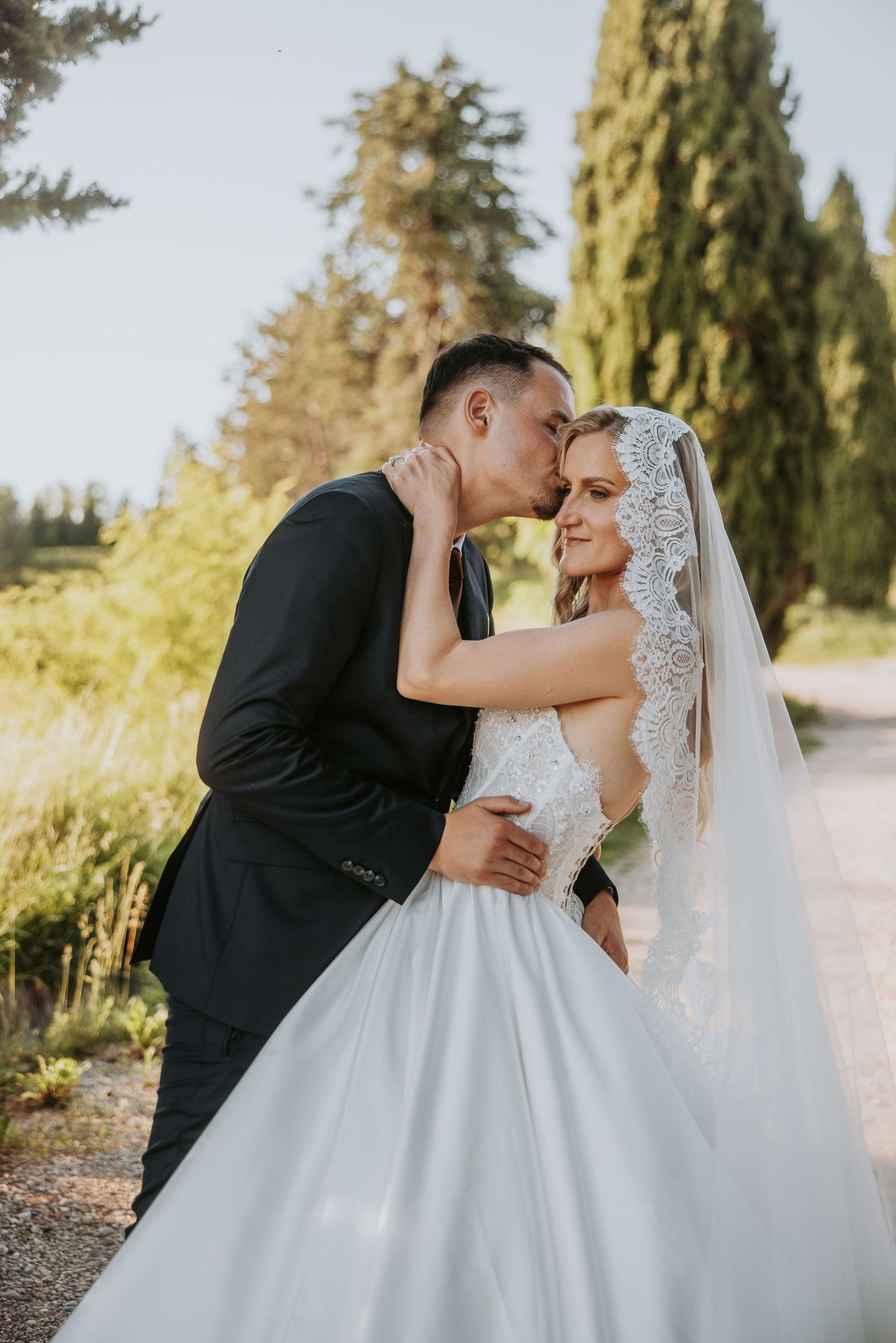 Groom in black suit tenderly kisses bride in white lace gown and veil outdoors.