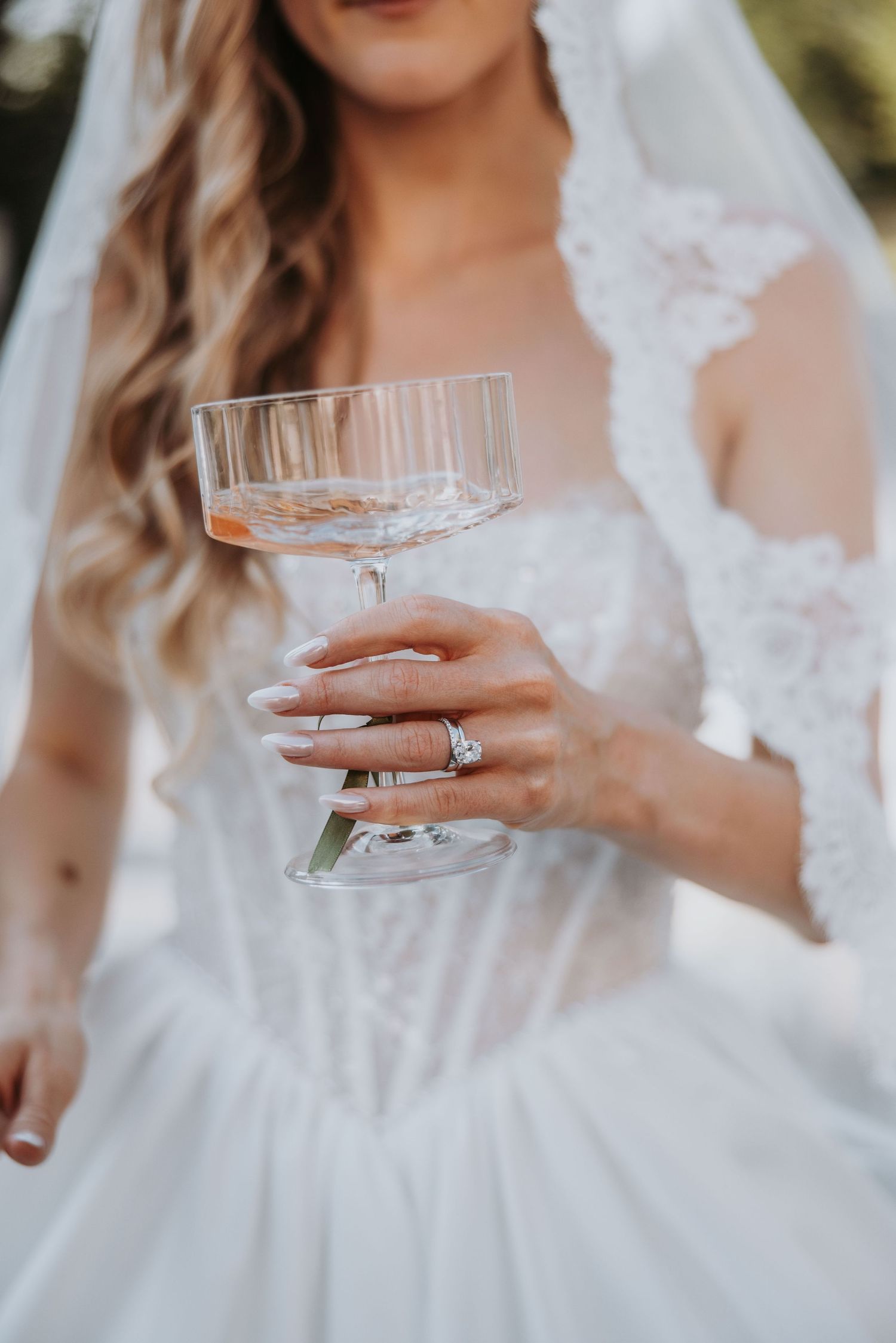 Bride in lace wedding dress holds champagne coupe glass showcasing diamond engagement and wedding rings.