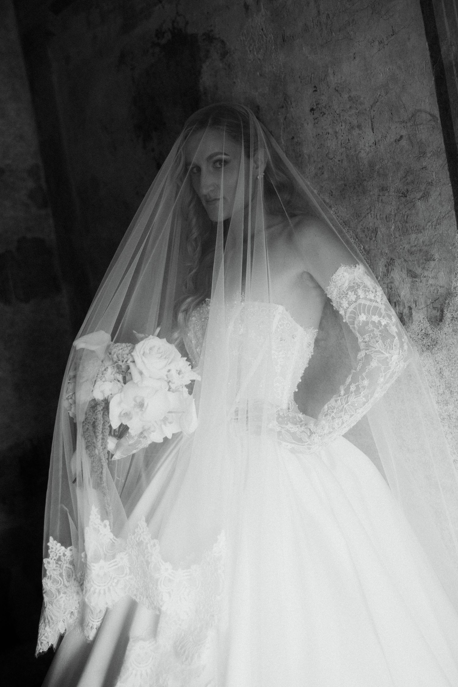 Black and white full-length portrait of a bride in a ball gown holding a bouquet beneath her lace veil.